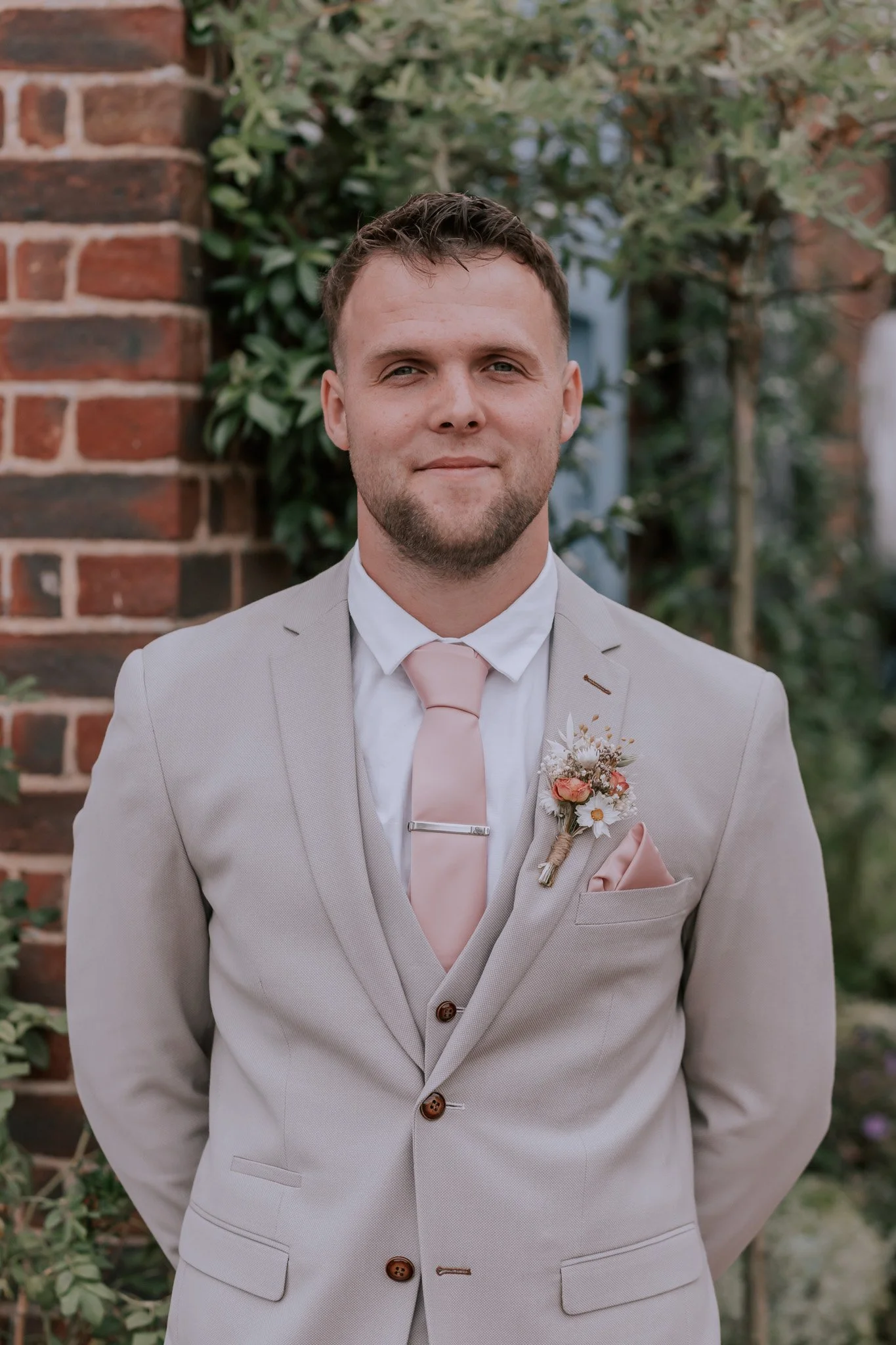 A man in a beige suit with a pink tie and boutonniere, standing outdoors in front of green foliage and a brick wall, facing the camera with a neutral expression.