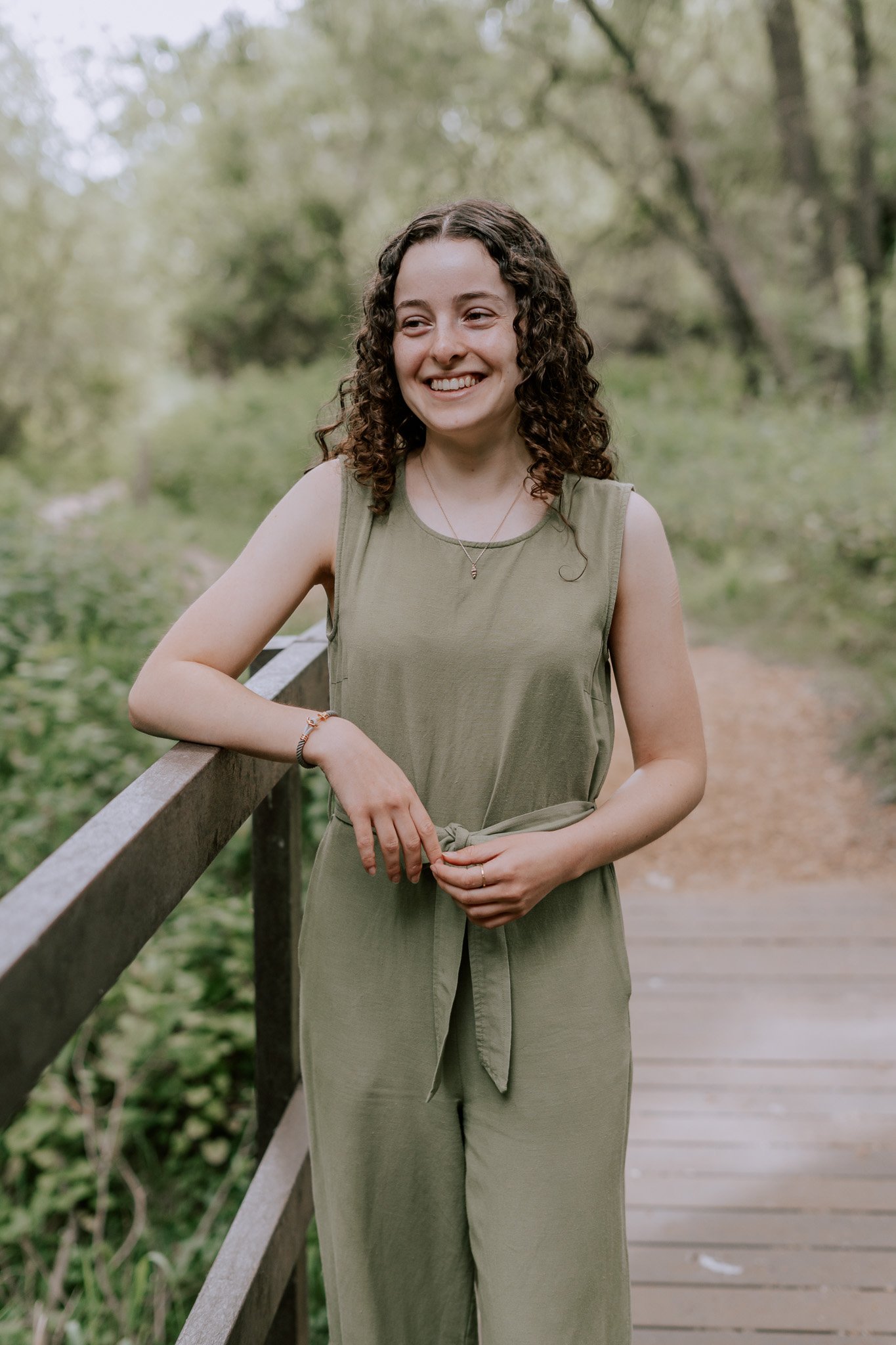 A young woman with curly brown hair smiling while standing on a wooden bridge in a green outdoor setting.