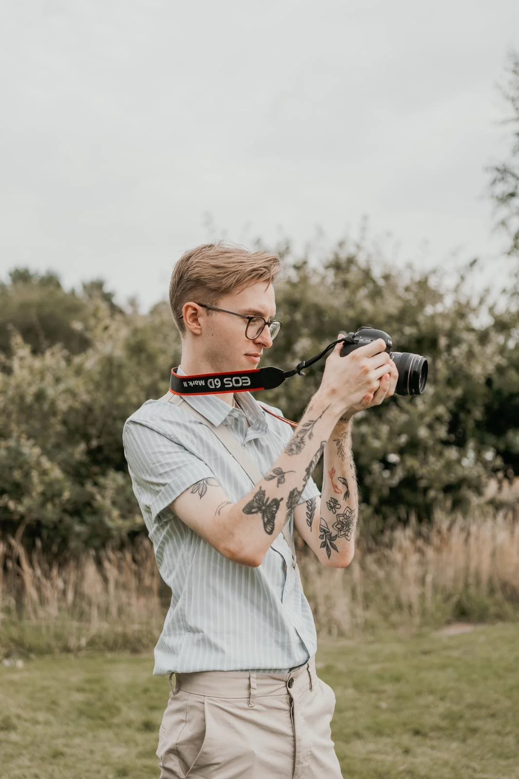 Young man with tattoos on arms and glasses taking a photo outdoors with a Canon camera.