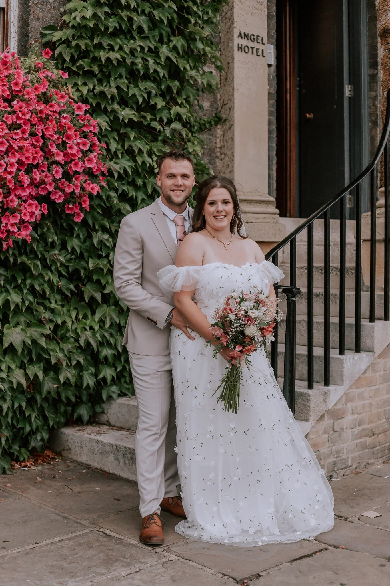 A newlywed couple standing on steps outside a building labeled 'Angel Hotel', surrounded by pink flowers and green ivy, smiling at the camera.