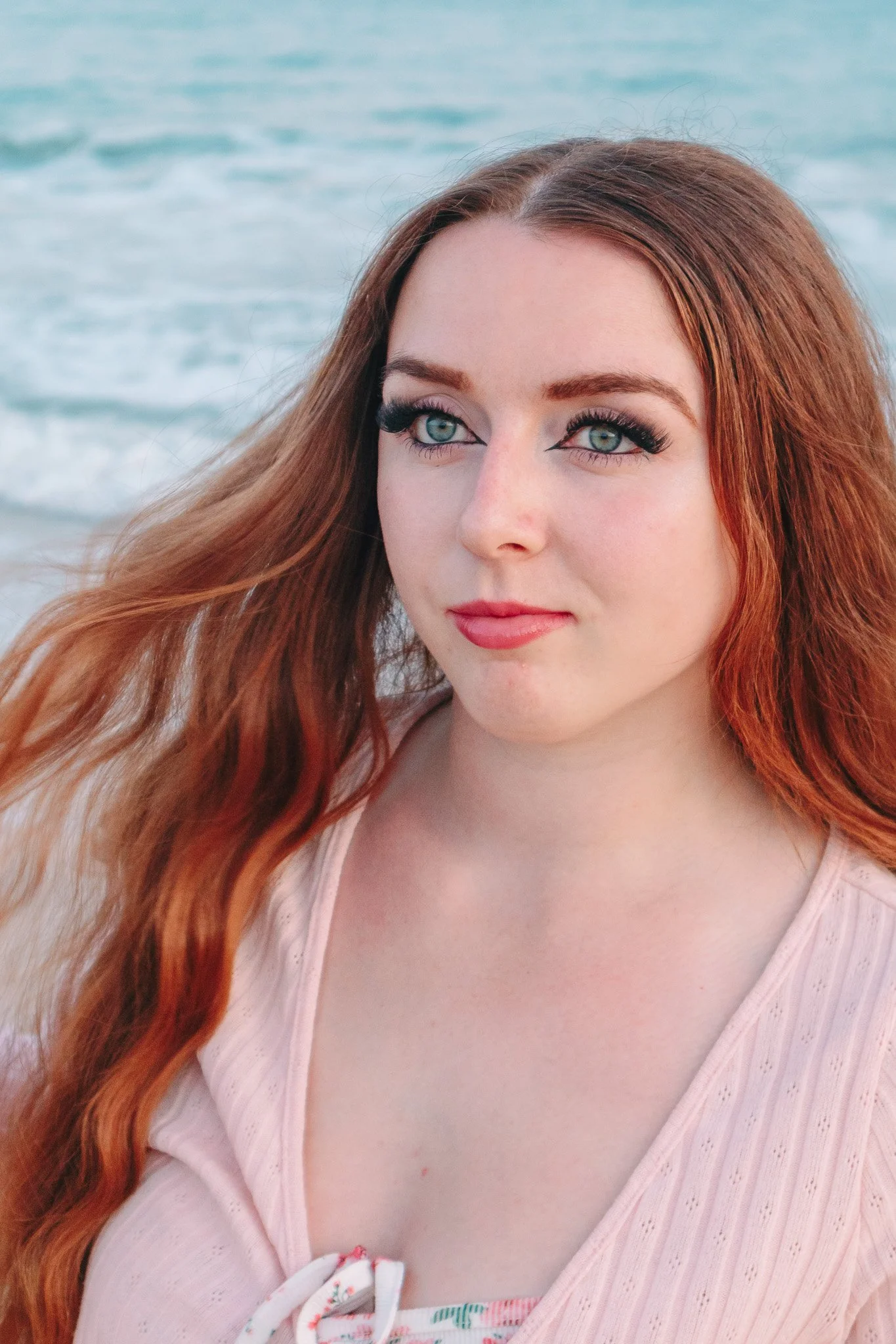 A woman with long, wavy red hair at the beach, looking slightly to the side.