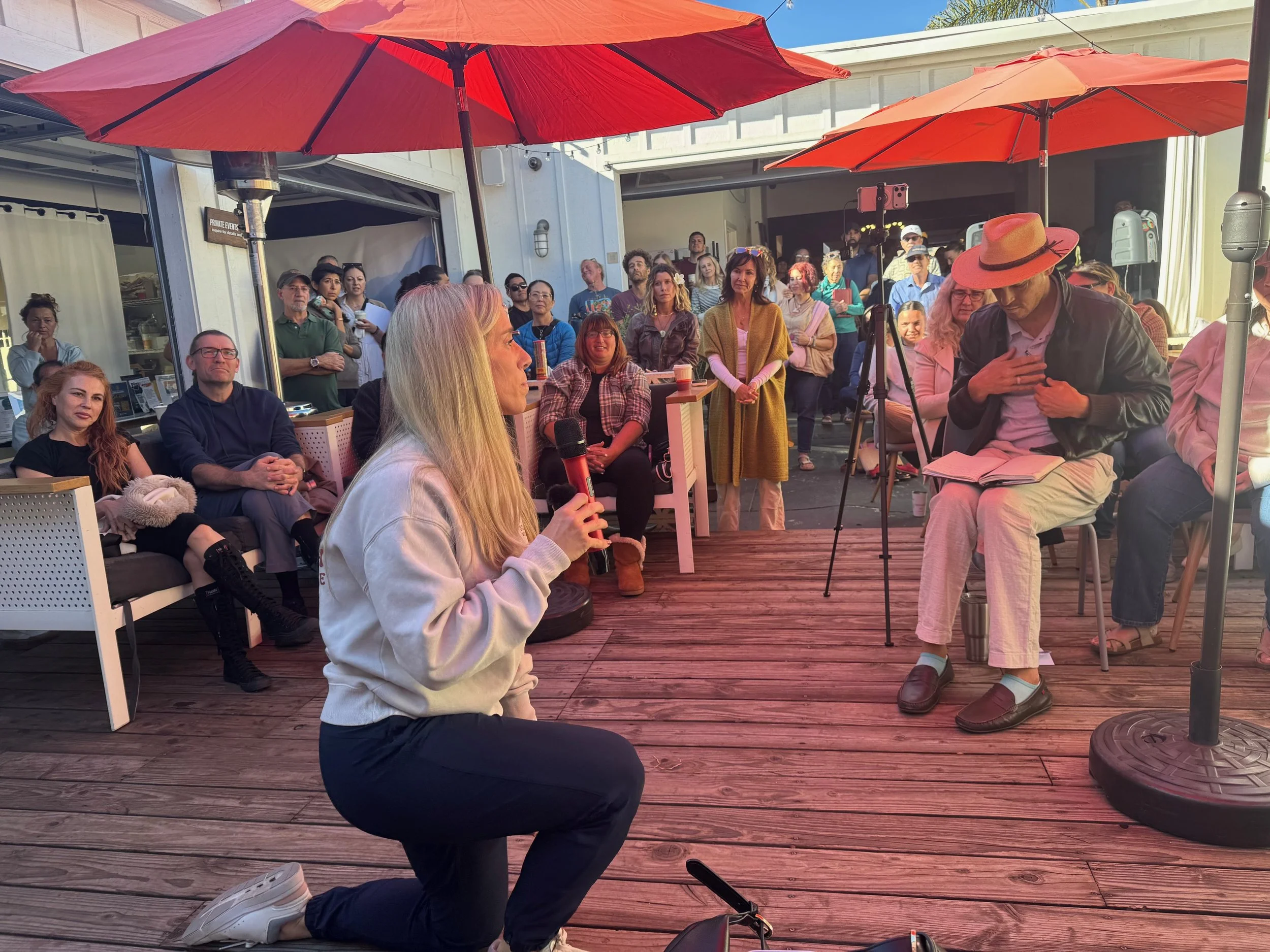 A woman with long blonde hair is kneeling and speaking into a microphone during an outdoor event under large red umbrellas. An audience is seated and standing around her, listening attentively. The setting appears casual with wooden flooring and people taking photos.