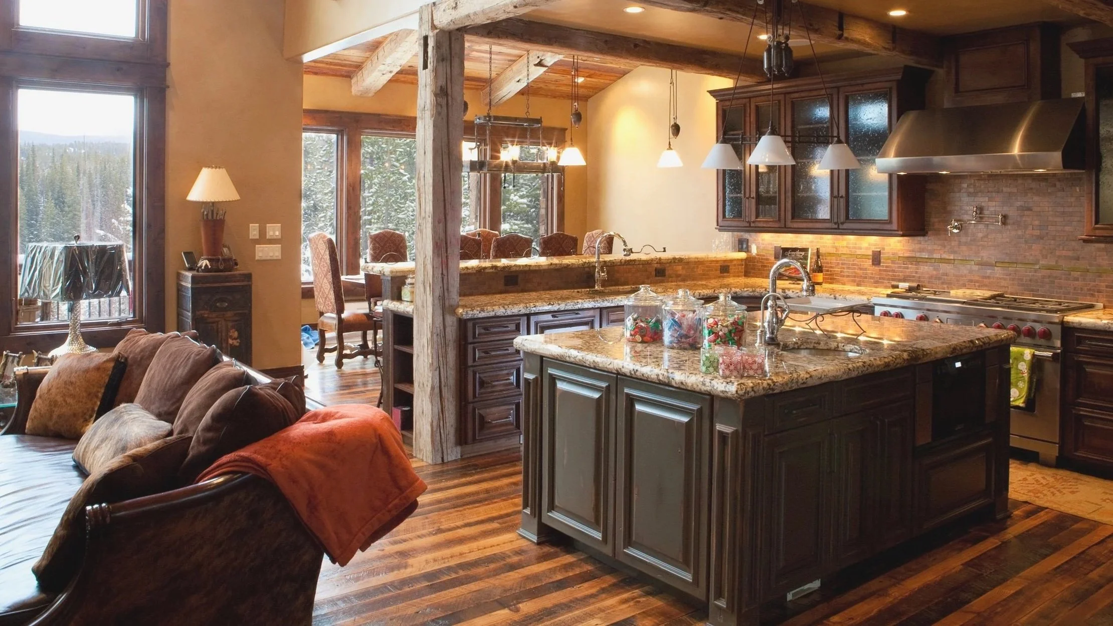 Interior view of a cozy, rustic-style kitchen with large windows showing snow-covered trees outside. The kitchen features wooden beams, dark wood cabinets, a granite island, and pendant lighting. A mounted deer head is on the wall above the windows, and a living area with chairs and a table is partially visible.