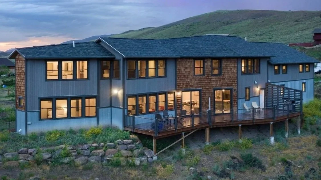 A modern two-story house with large windows and a wooden deck overlooking a rural landscape at dusk.