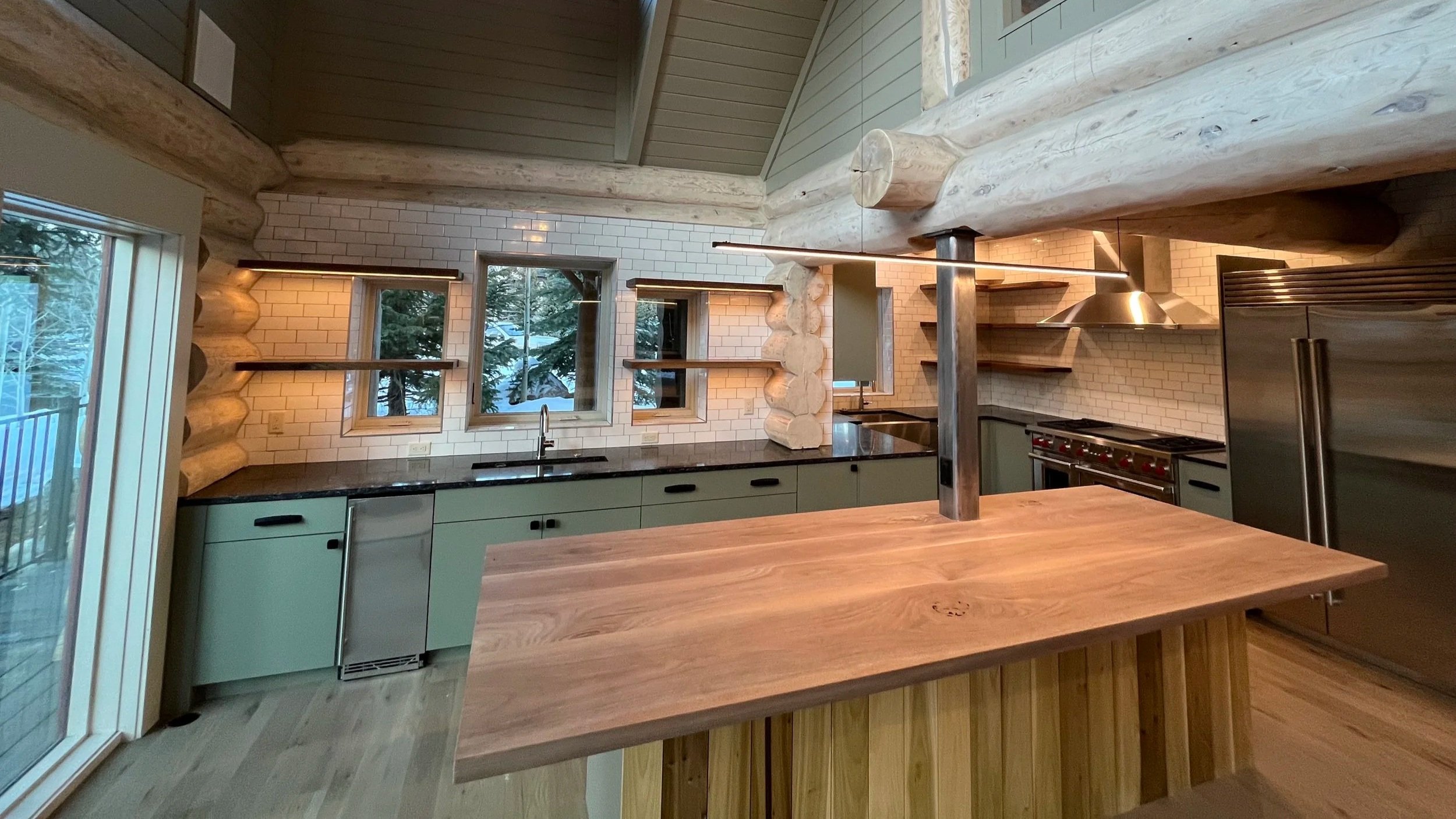Interior view of a modern kitchen with white brick walls, black countertops, and mint green cabinets. There are large windows and a skylight, with visible wooden beams and a wooden island in the foreground.