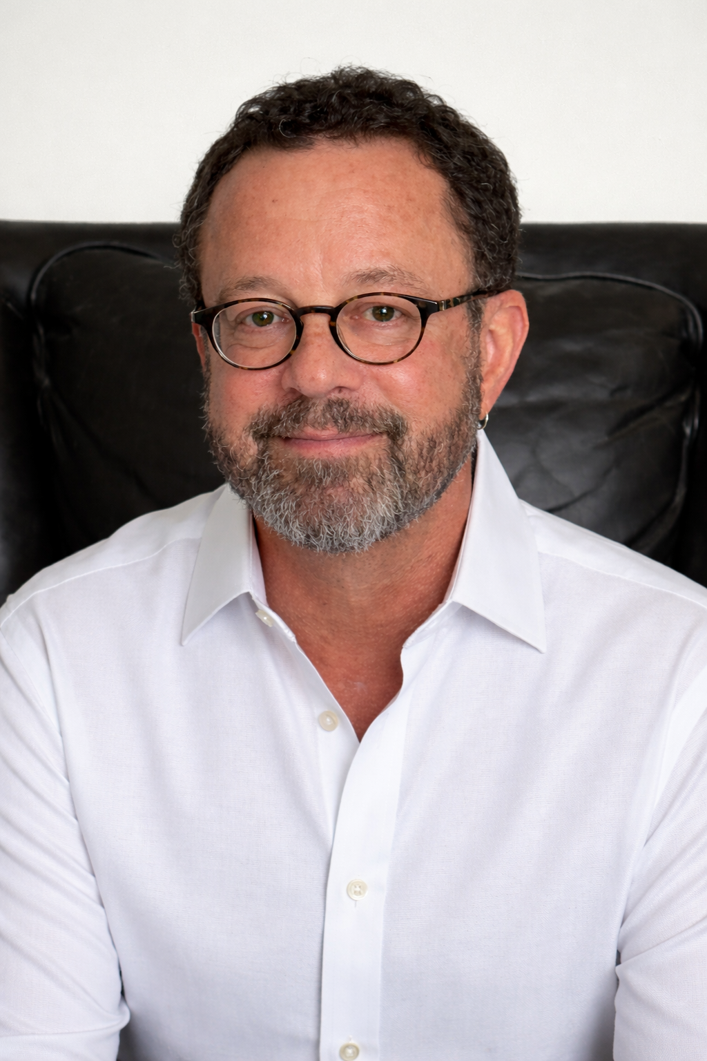 A middle-aged man with curly dark hair, glasses, and a beard, dressed in a white button-up shirt, sitting on a black leather chair with a plain white wall background.