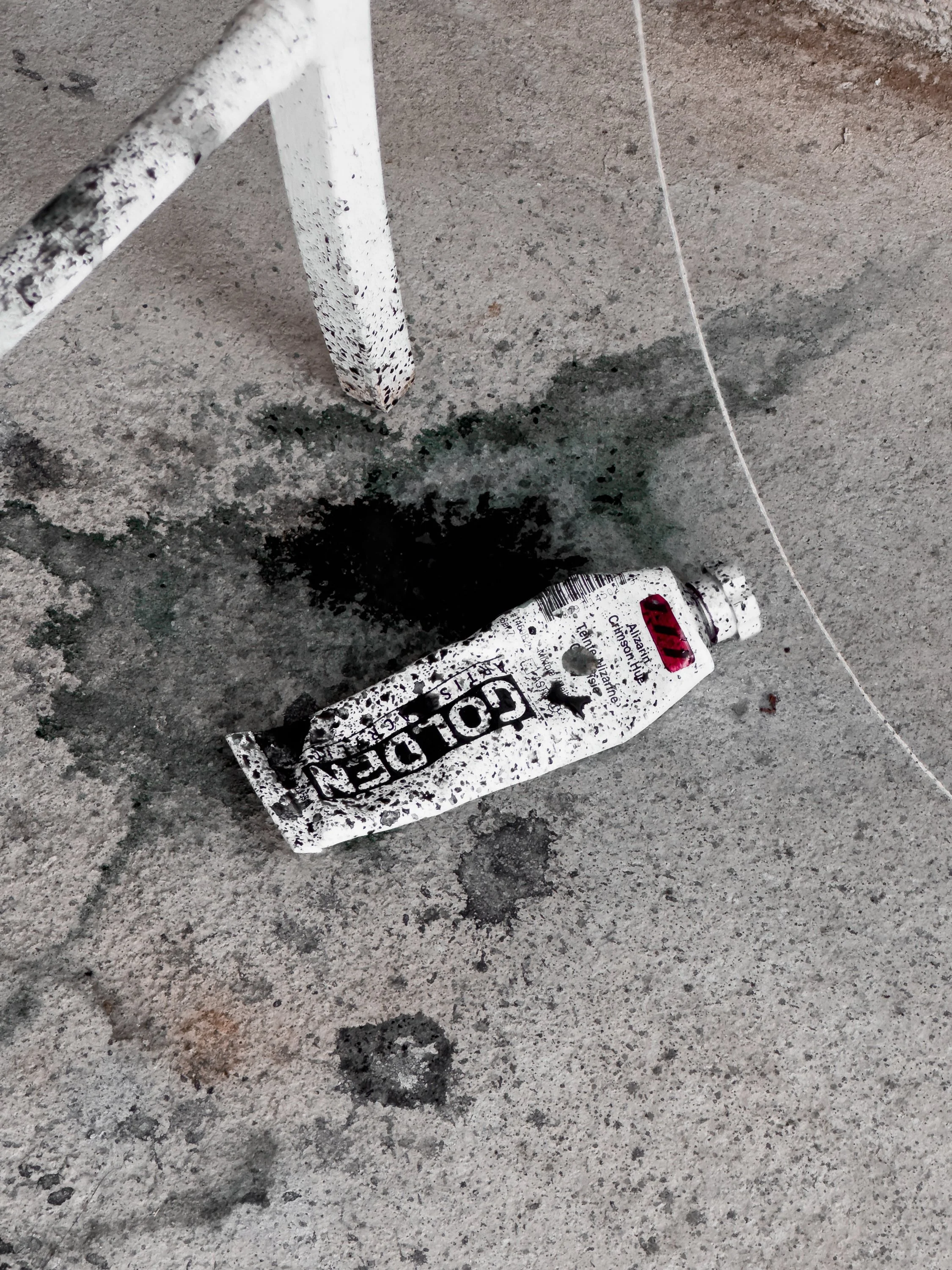 A crumpled tube of paint labeled 'GOLDEN' lies on a concrete floor, with black paint spilled around it and a white metal chair leg nearby.