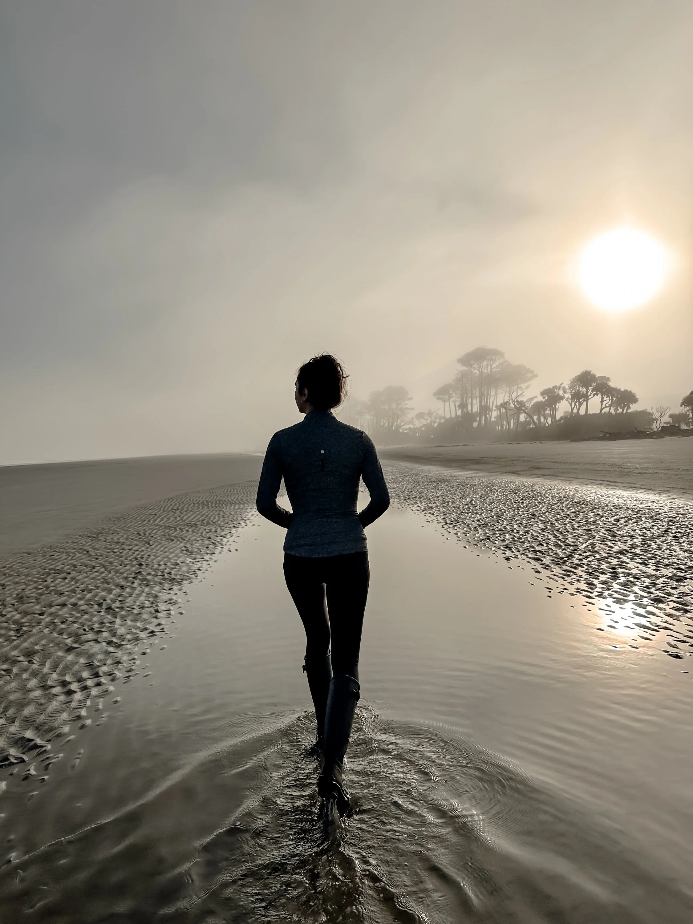 A person walking on a beach at sunset, with trees in the distance and a cloudy sky.