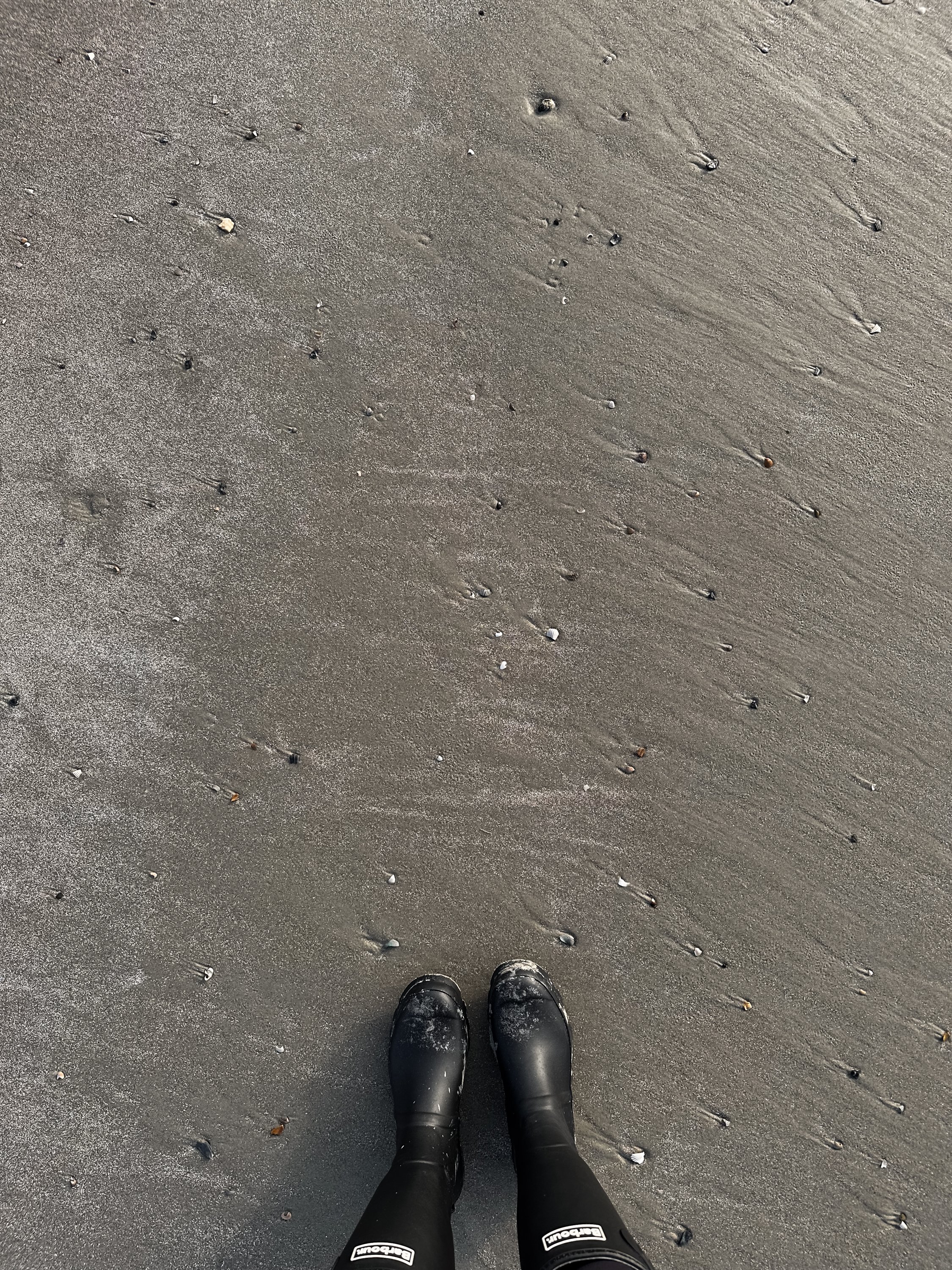 View of sandy beach with small pebbles, boots worn by person standing on the sand.