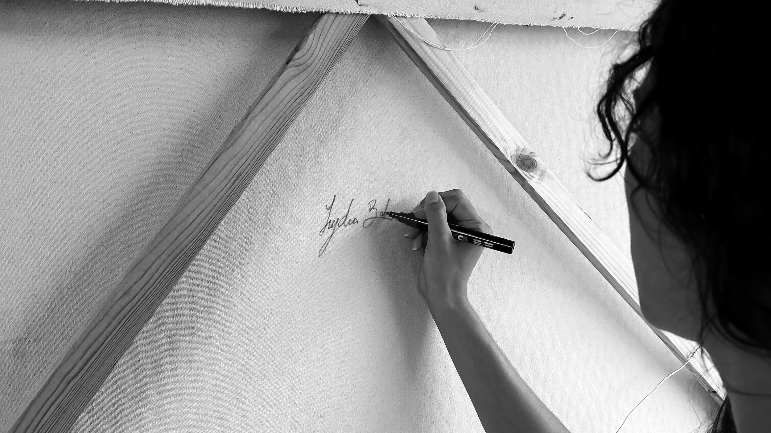 A person with curly hair writing a signature on a wall or canvas with a black marker, with a wooden triangle frame above.