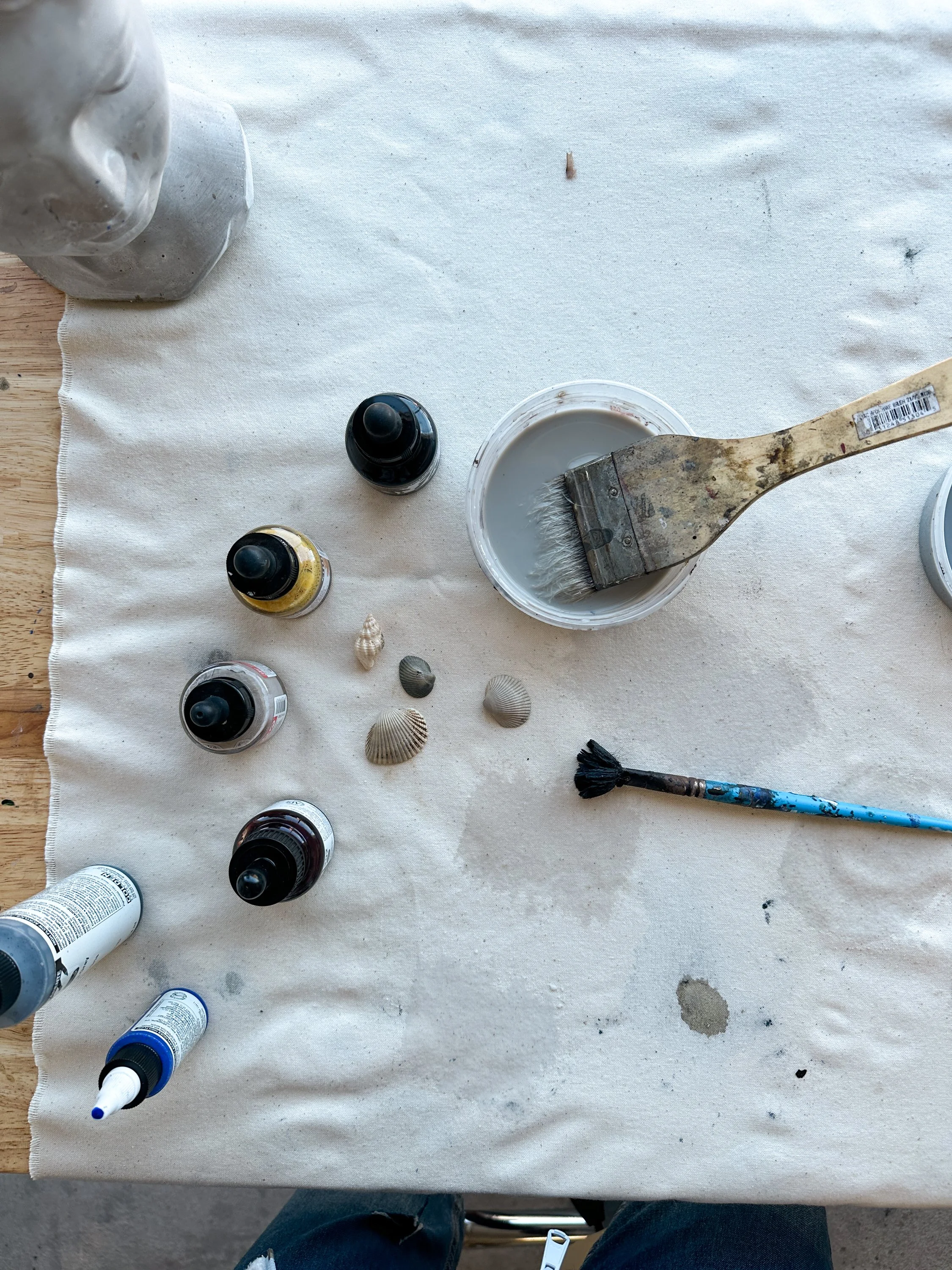 Paint supplies on a white cloth-covered table, including bottles of paint, seashells, a paintbrush in a container of grey paint, and a cleaning brush.