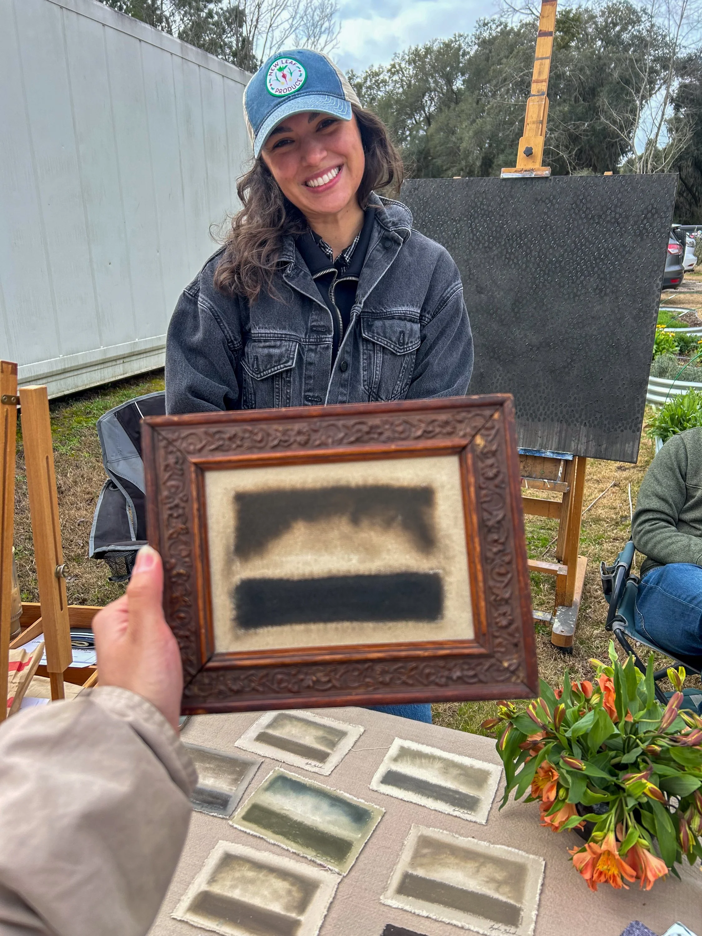 A woman with long wavy hair smiling, wearing a denim jacket and a baseball cap with 'New Leaf Produce' logo, standing outdoors near a gray wall and trees. In front of her, an outdoor display of framed artwork and small rectangular tiles on a table.