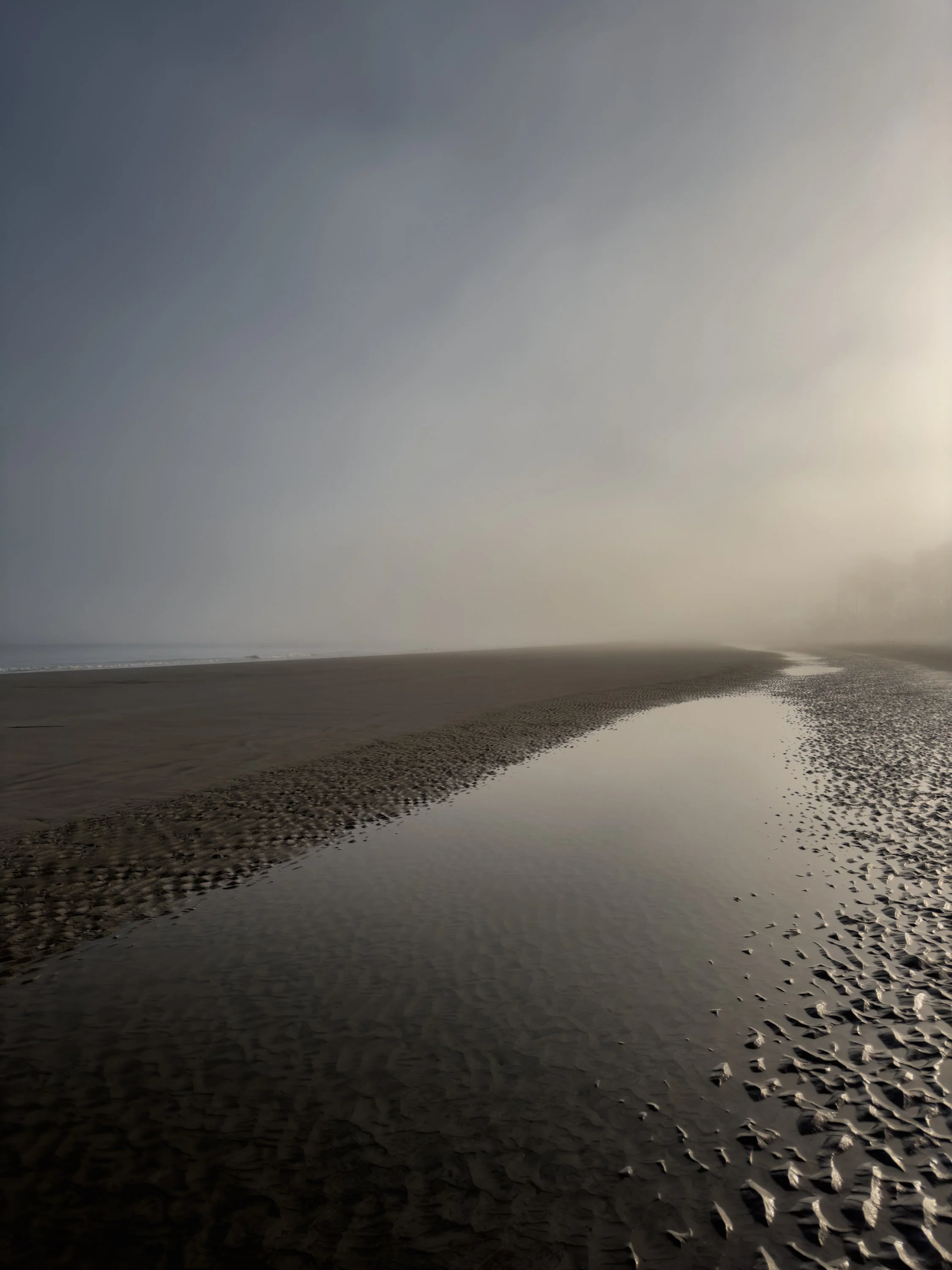 A beach scene with sand, a narrow water stream, and a foggy sky.