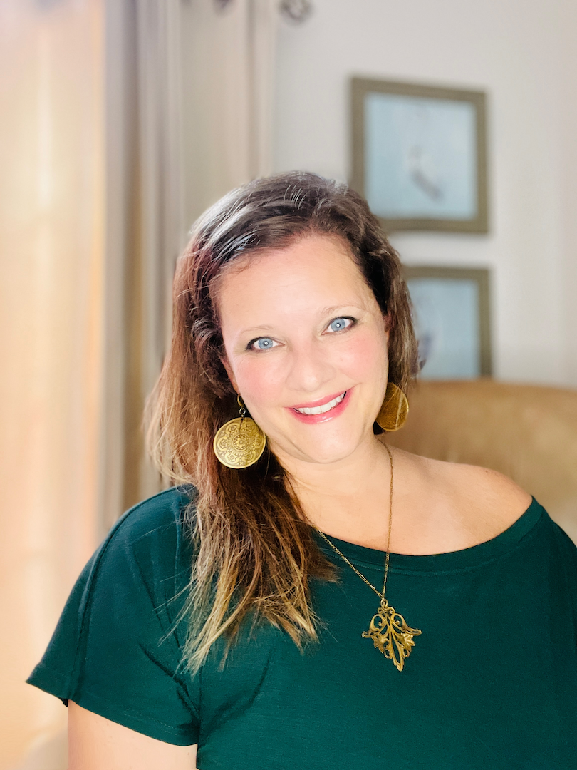 A smiling woman with blue eyes and brown hair wearing gold earrings and a gold leaf necklace, sitting in a room with framed pictures on the wall.