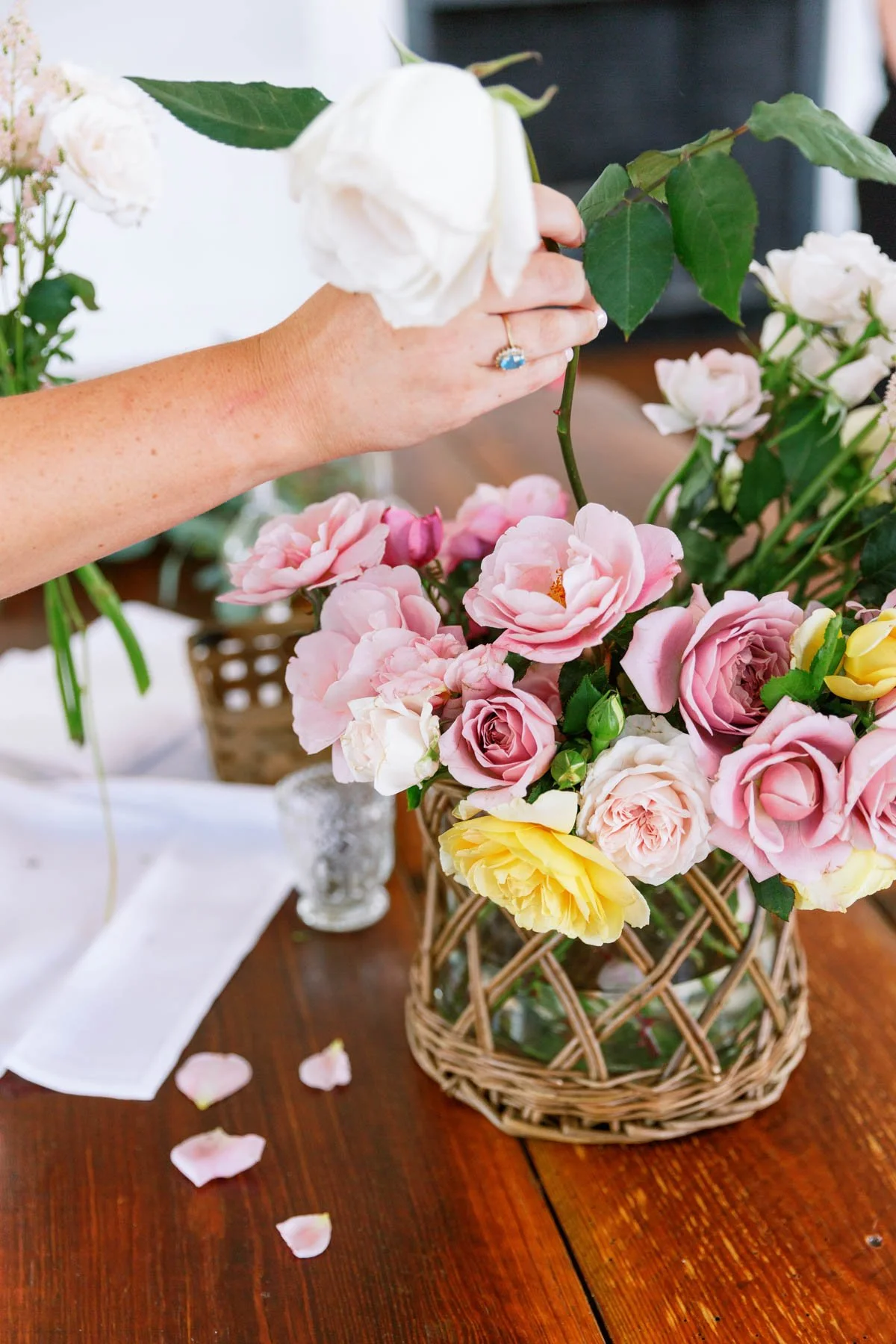 lose-up of a hand holding a white rose above pink flowers on a table.