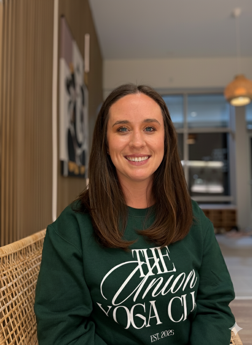 A smiling woman with shoulder-length curly brown hair wearing a light blue V-neck T-shirt, standing against a wooden-paneled wall.
