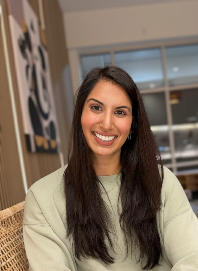 A woman with long, wavy brown hair smiling and crossing her arms against a wooden wall.