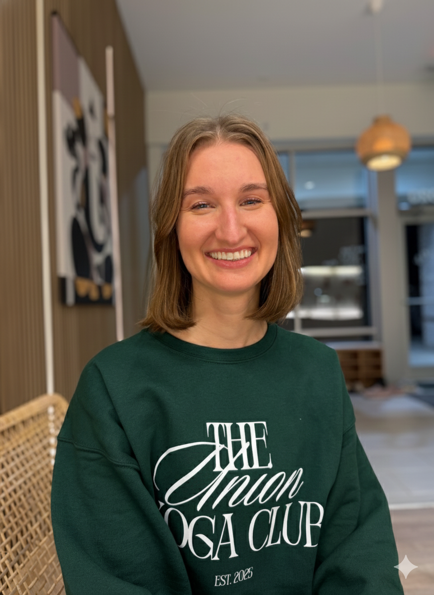 Smiling woman with shoulder-length brown hair wearing a light gray sweater standing against a wooden panel wall.
