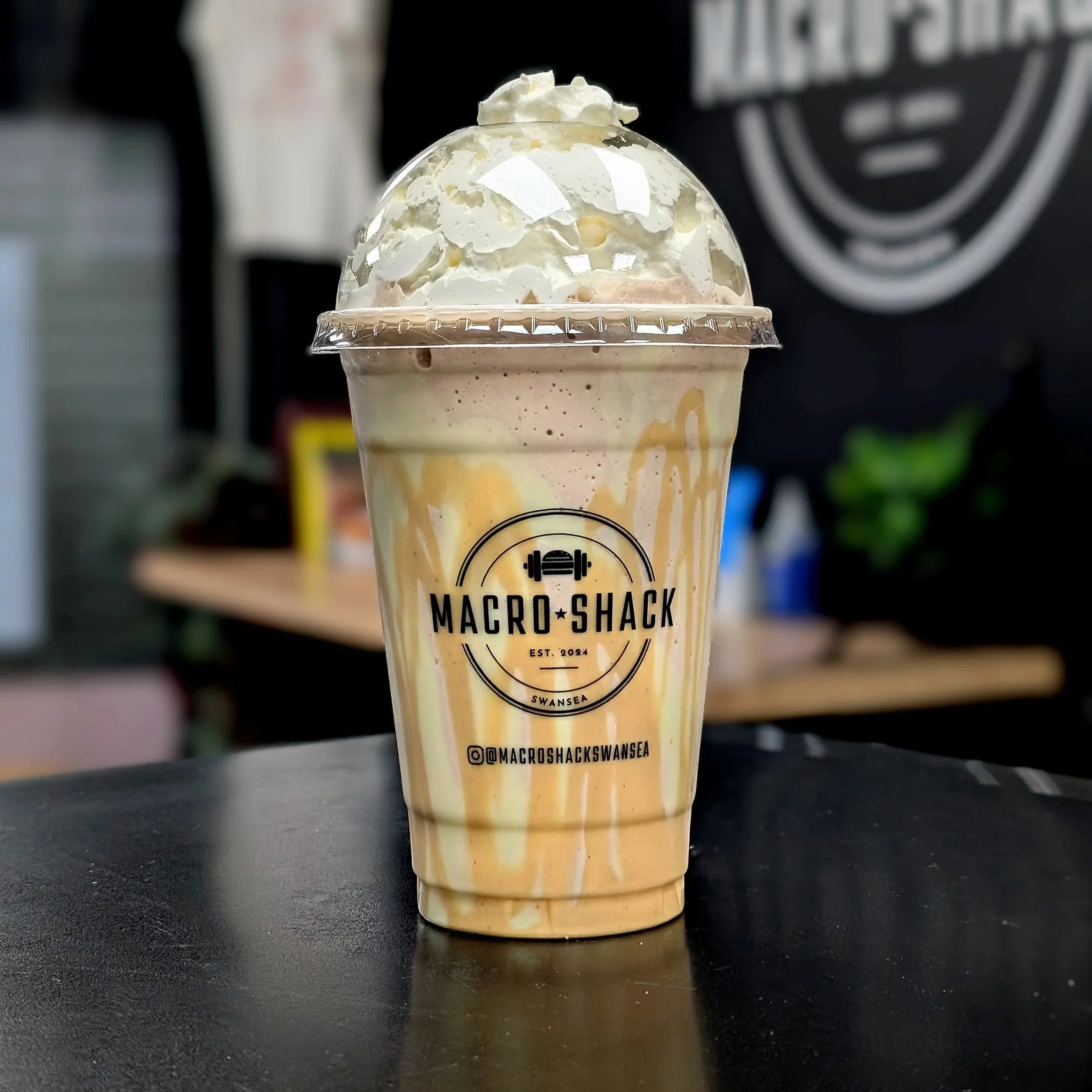 A cold high protein milkshake with whipped cream and caramel drizzle in a clear plastic cup, featuring the Macro Shack logo and social media handle, placed on a dark surface with a blurred background.