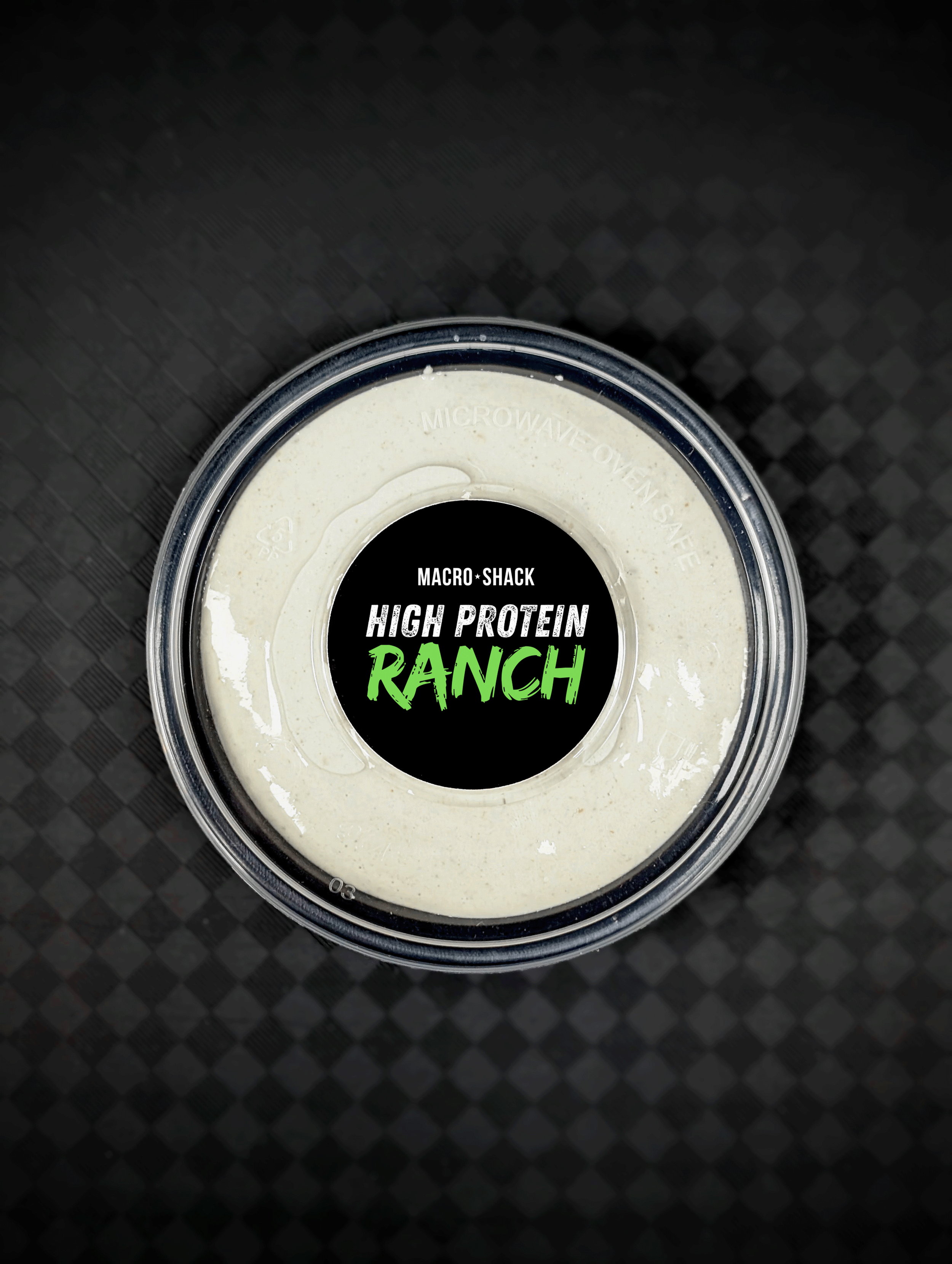 Top view of a container of high protein ranch dip with a black label featuring green and white text on a dark checkered surface.
