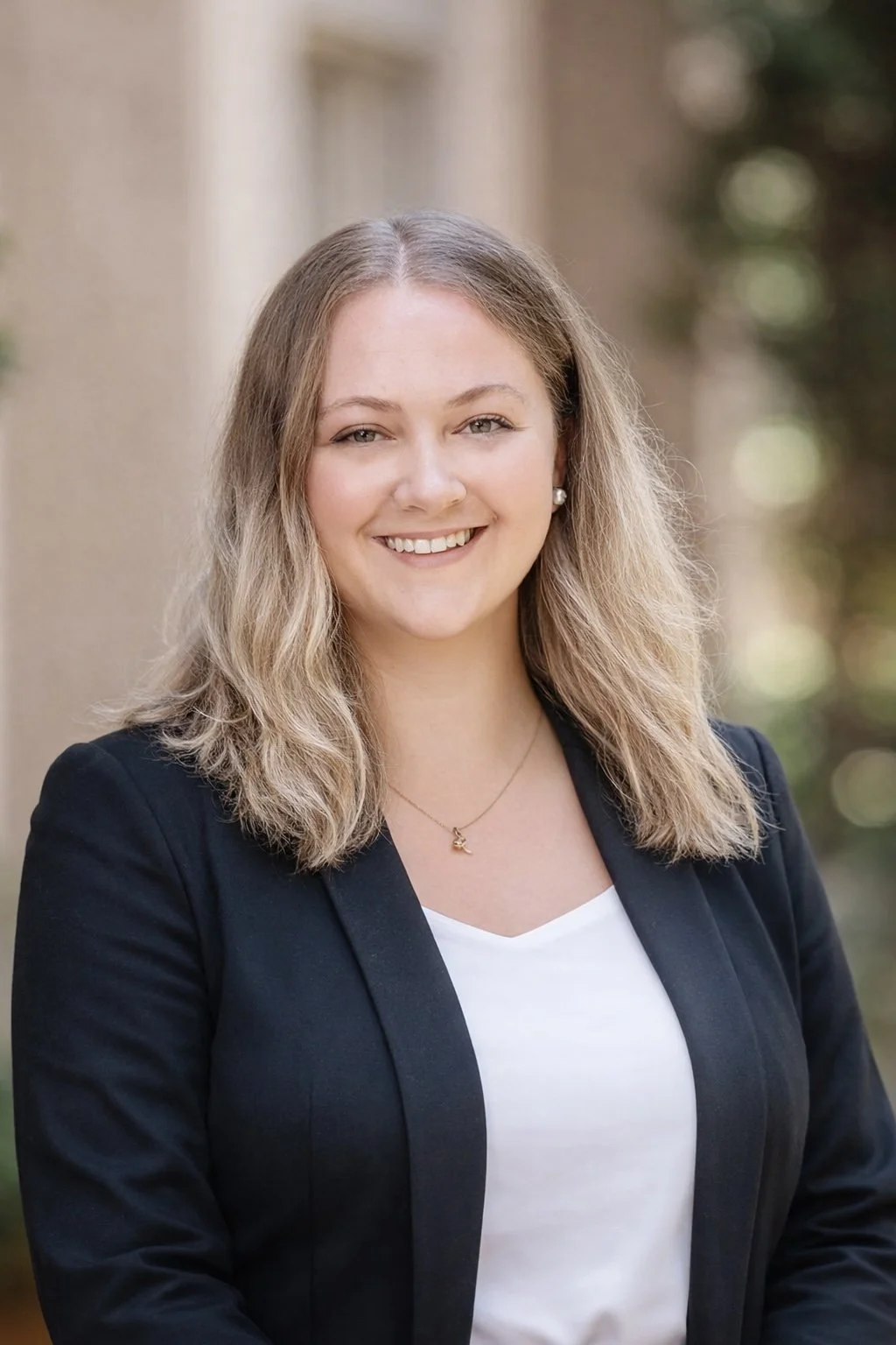 A young woman with blonde wavy hair, wearing a black blazer, white top, pearl earrings, and a gold necklace, smiling outdoors with blurred trees and wall background.