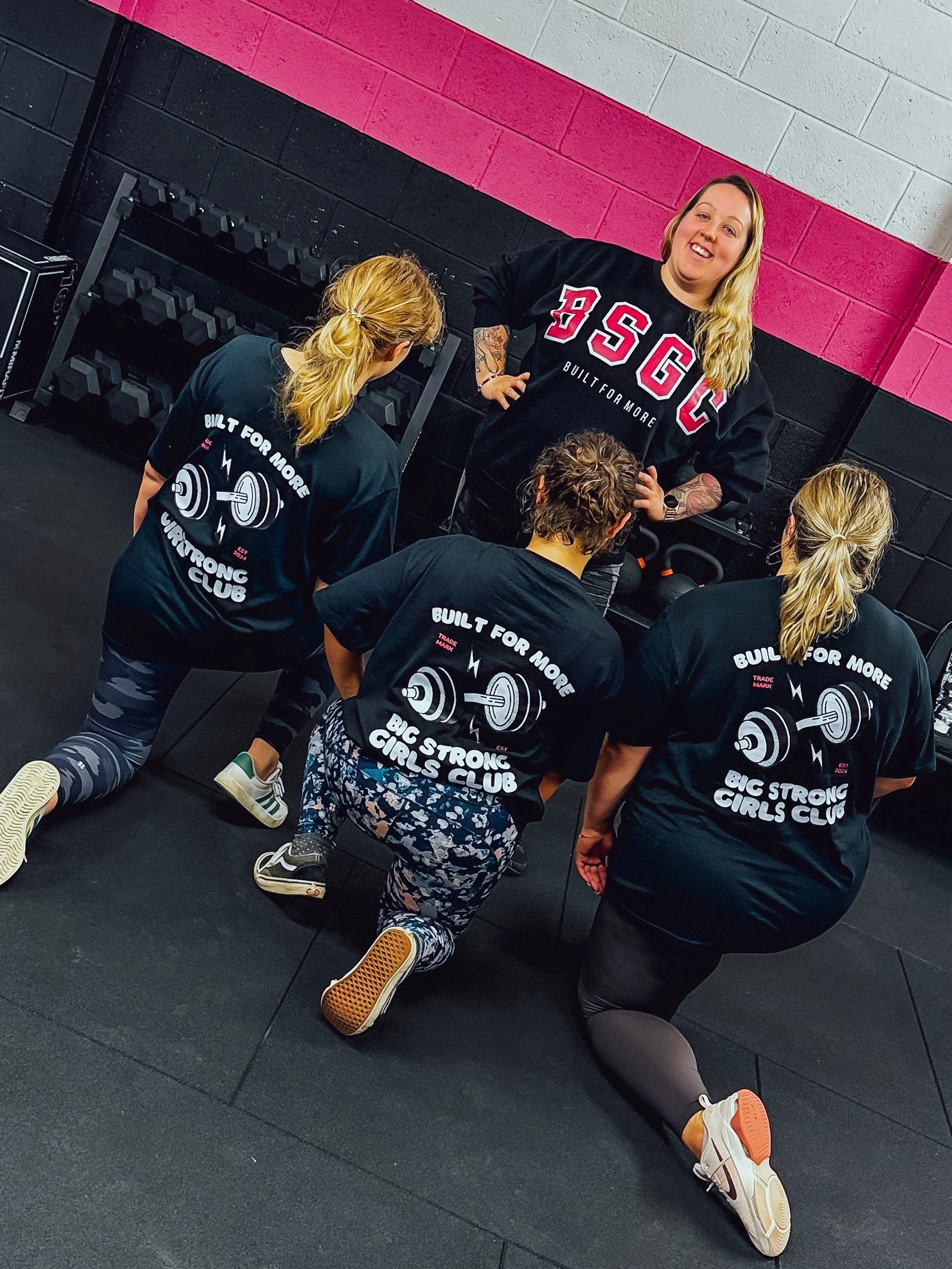 Group fitness class with women kneeling on the floor in front of a female instructor, all wearing matching black T-shirts with a weightlifting graphic and the text 'Built for More Big Strong Girls Club'. They are in a gym with pink and black walls and a standard set of dumbbells on a rack.