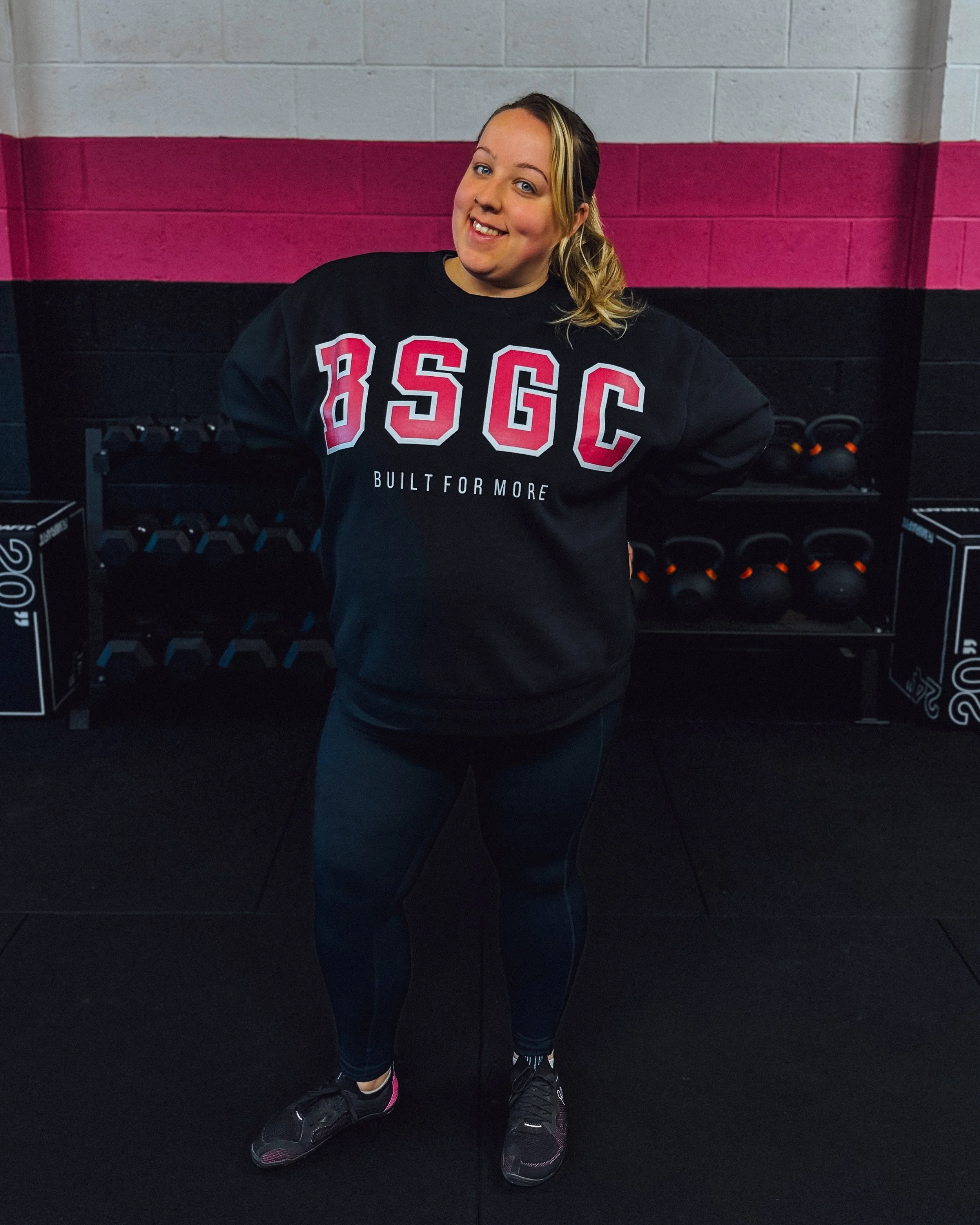 A woman standing in a gym, wearing a black sweatshirt with red and white lettering 'BSGC' and the slogan 'Built For More,' black leggings, and black sneakers, with gym equipment like kettlebells and dumbbells in the background.