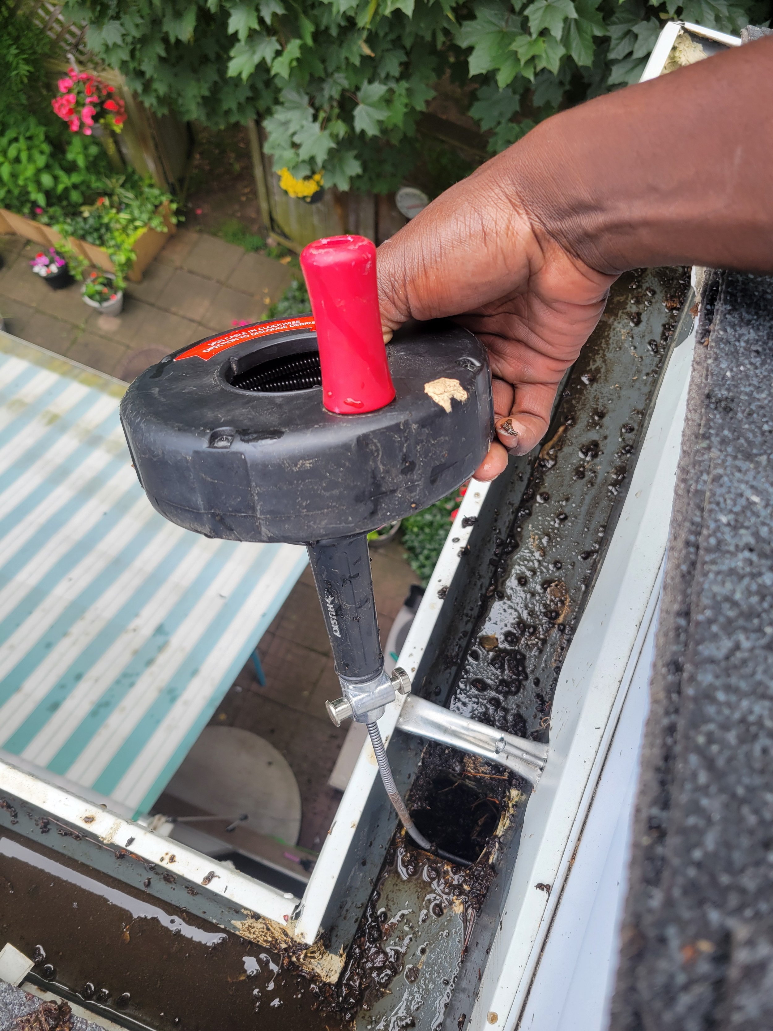 A person working on cleaning or repairing a gutter on a roof, with tools and debris inside the gutter, above a backyard with potted plants and a striped canopy.