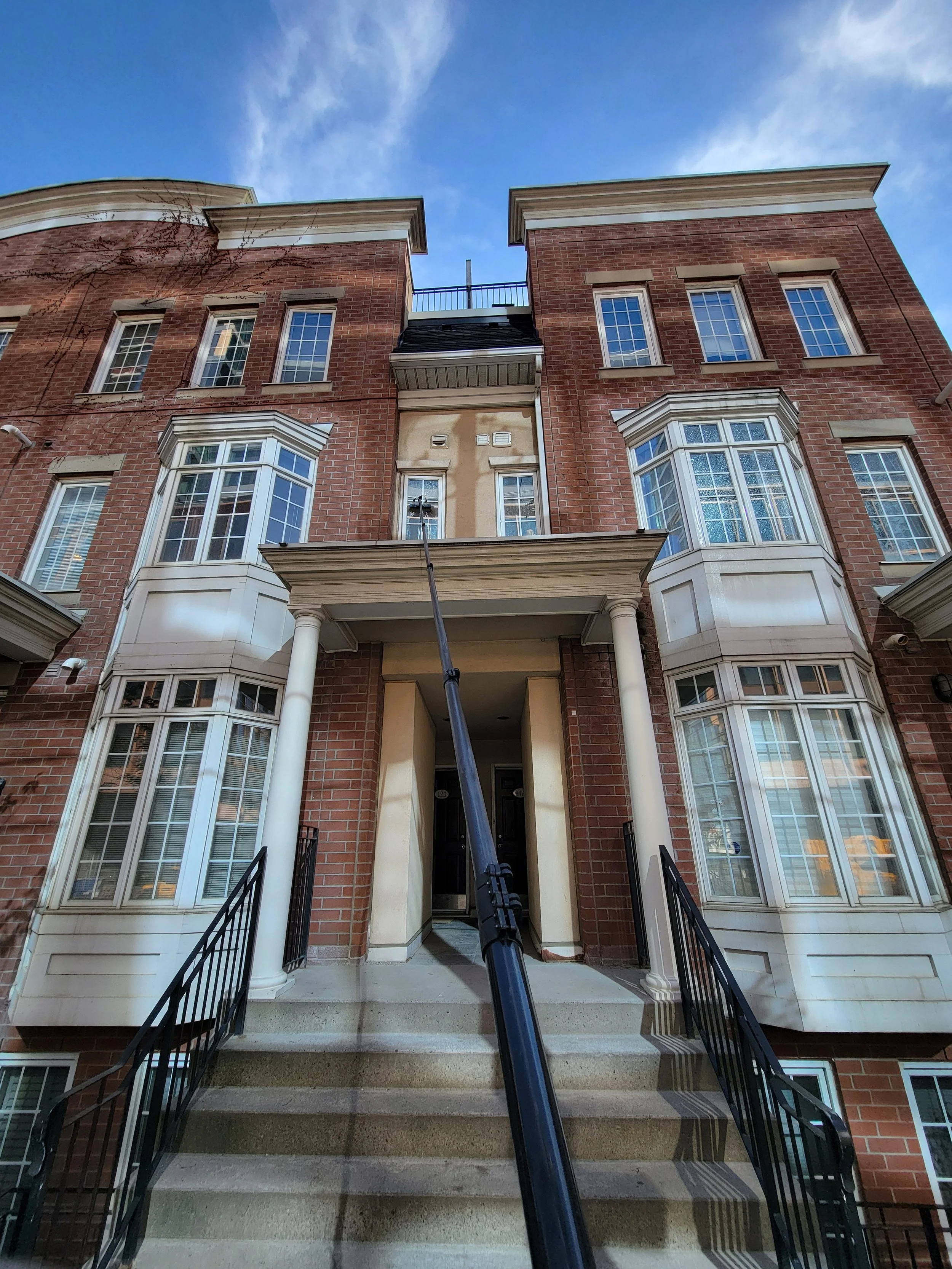 Photo of a multi-story brick residential building with front stairs, columns, and large bay windows, taken from ground level looking upward.