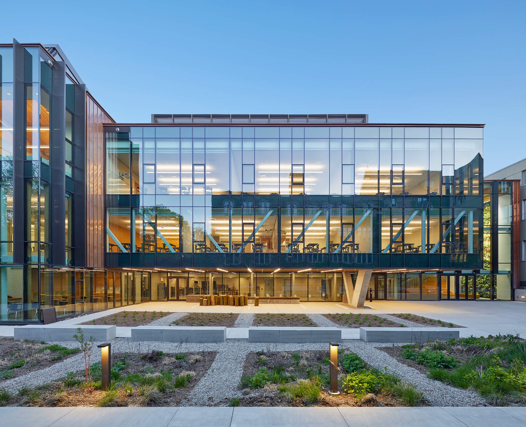 Modern multi-story glass building with illuminated interior, outdoor landscaping, and pathway, during evening with a clear blue sky.