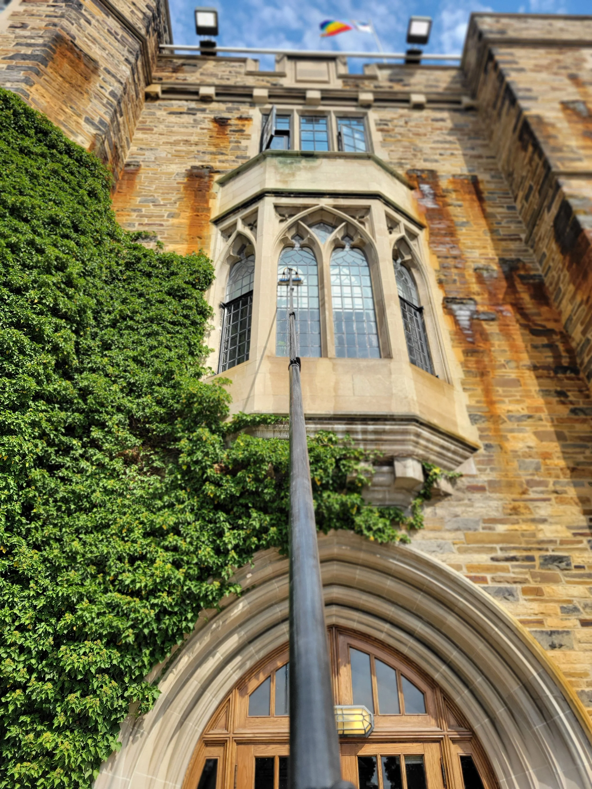 Low-angle view of a historic brick building with large arched windows, a wooden doorway, and a flag flying on the roof. There's greenery growing on the side of the building.