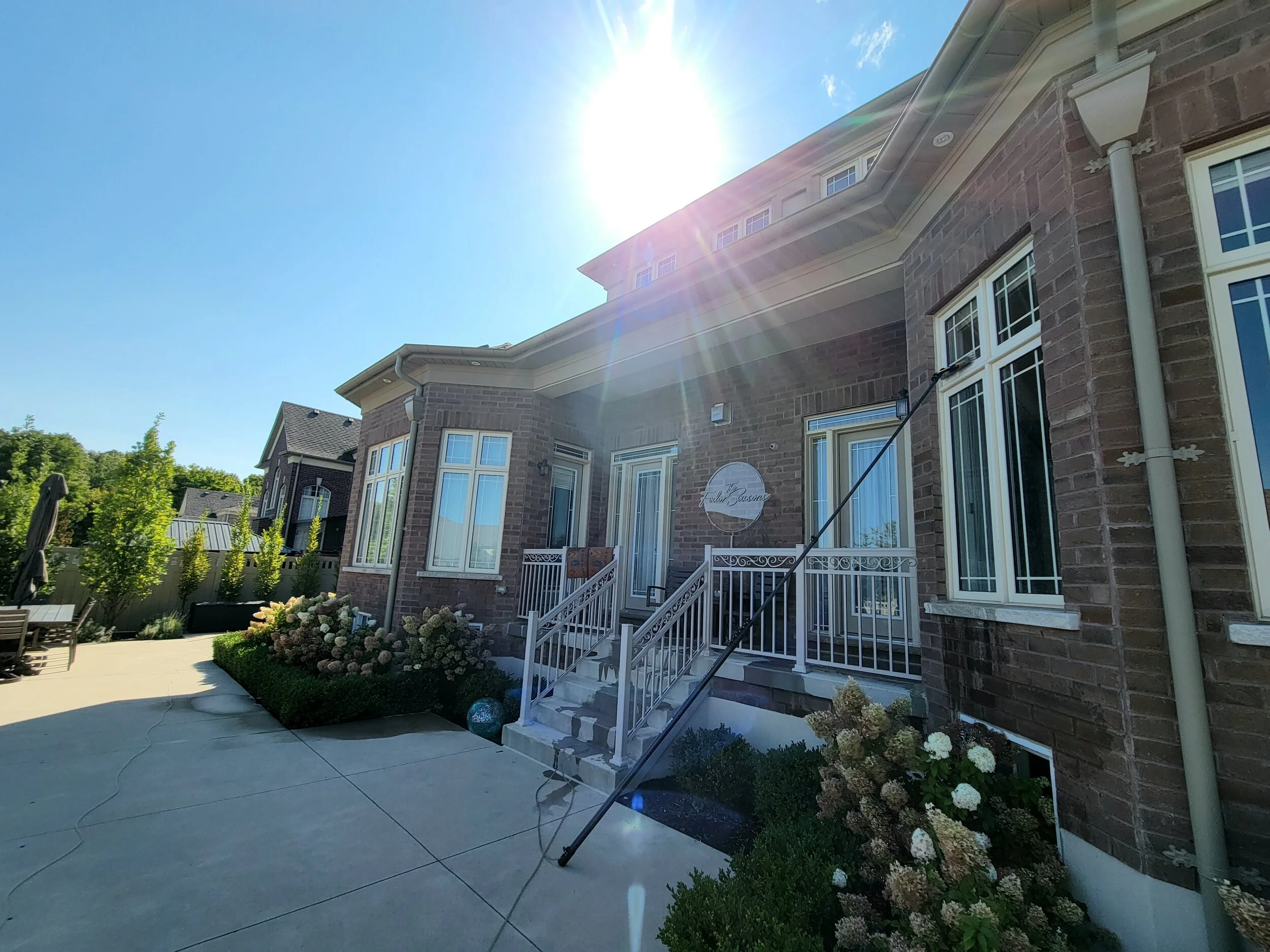 A brick house with large windows and a small porch with white railings, surrounded by manicured bushes and flowers under a bright sunny sky.