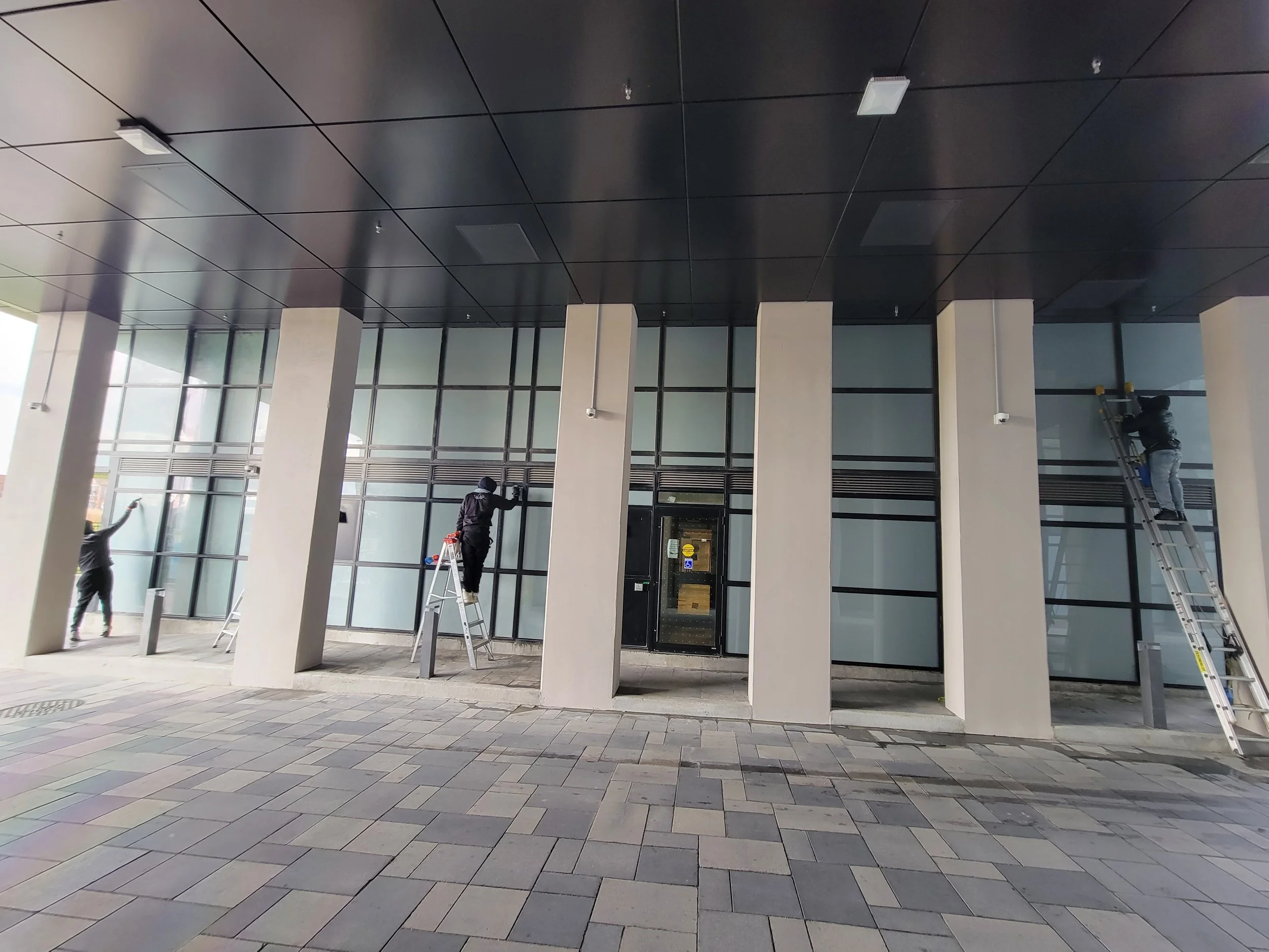 Three workers on ladders cleaning large glass windows in a modern building exterior.