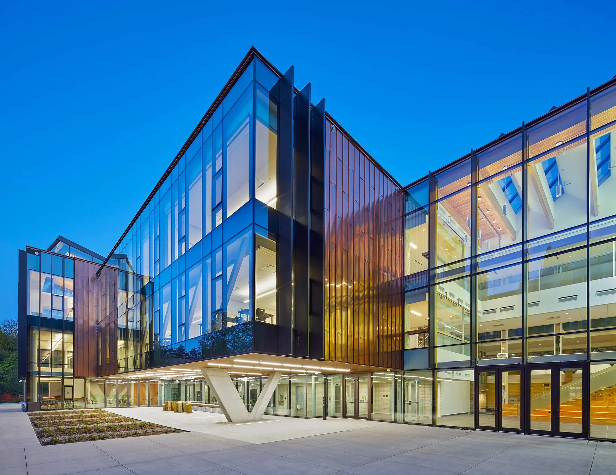 Modern glass building with illuminated interior, triangular support structure, and landscaped walkway at dusk.