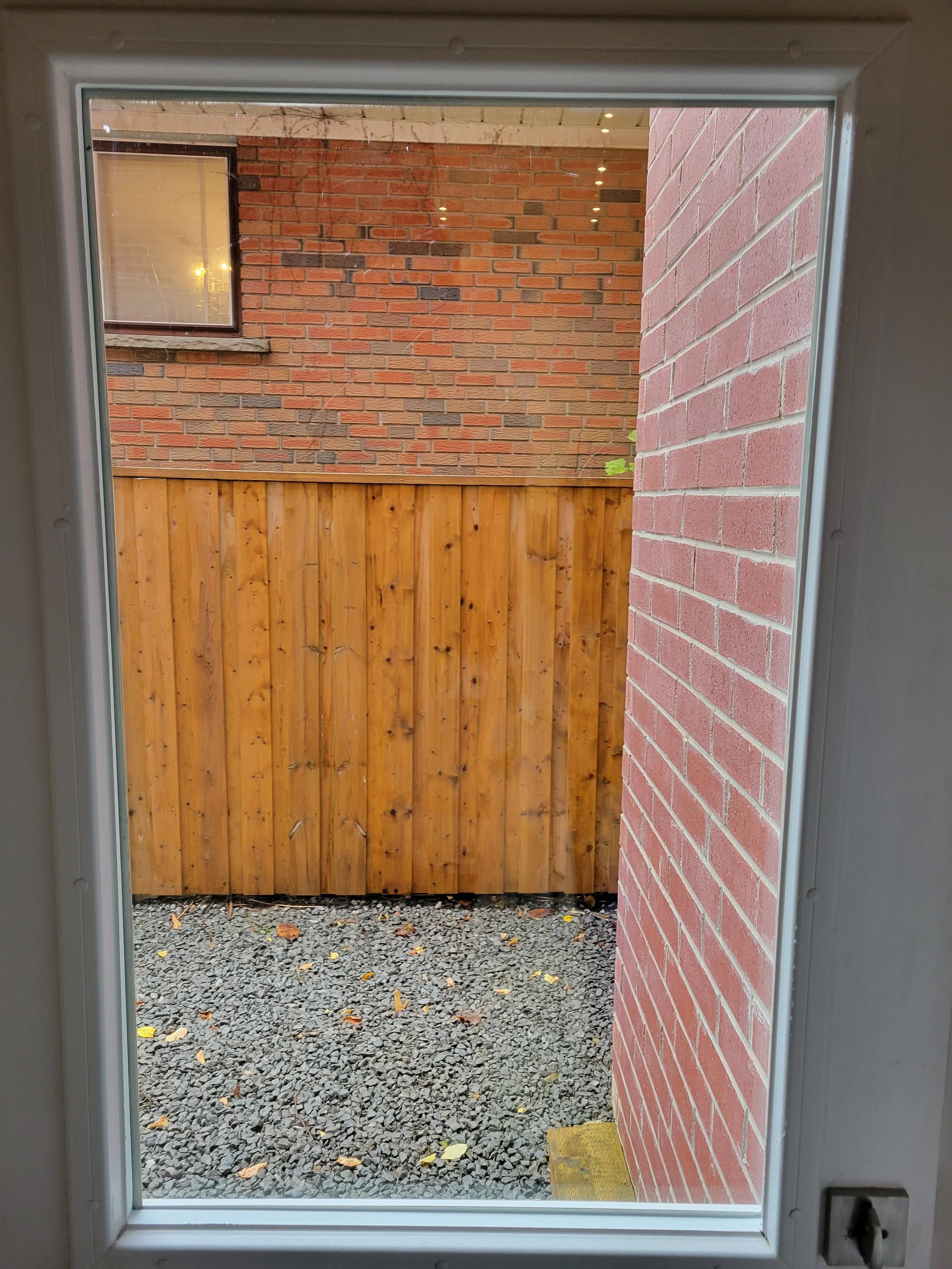 View through a window showing a small outdoor area with a gravel ground, a wooden fence, and brick walls of neighboring buildings.