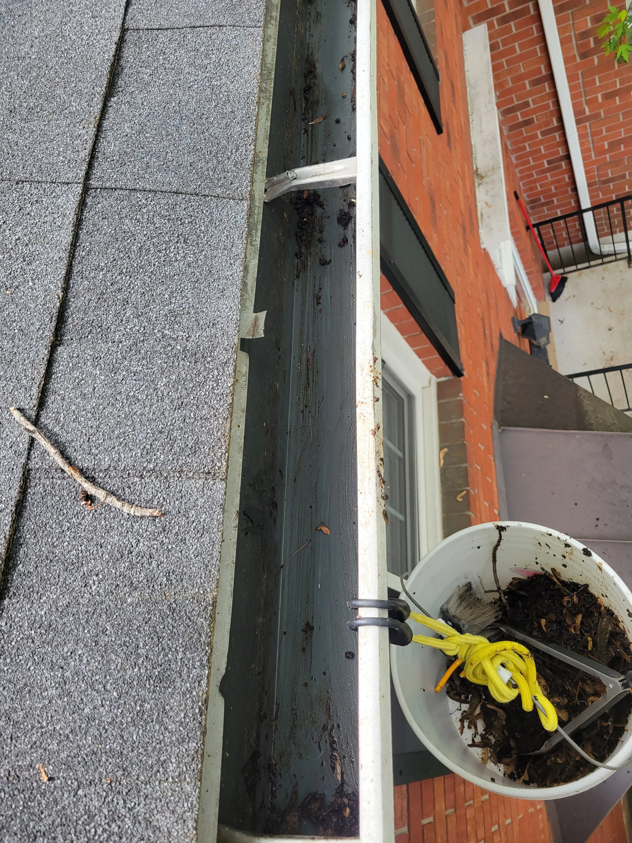 Roof gutter filled with leaves and debris, with a bucket of collected debris hanging on the edge of the roof.