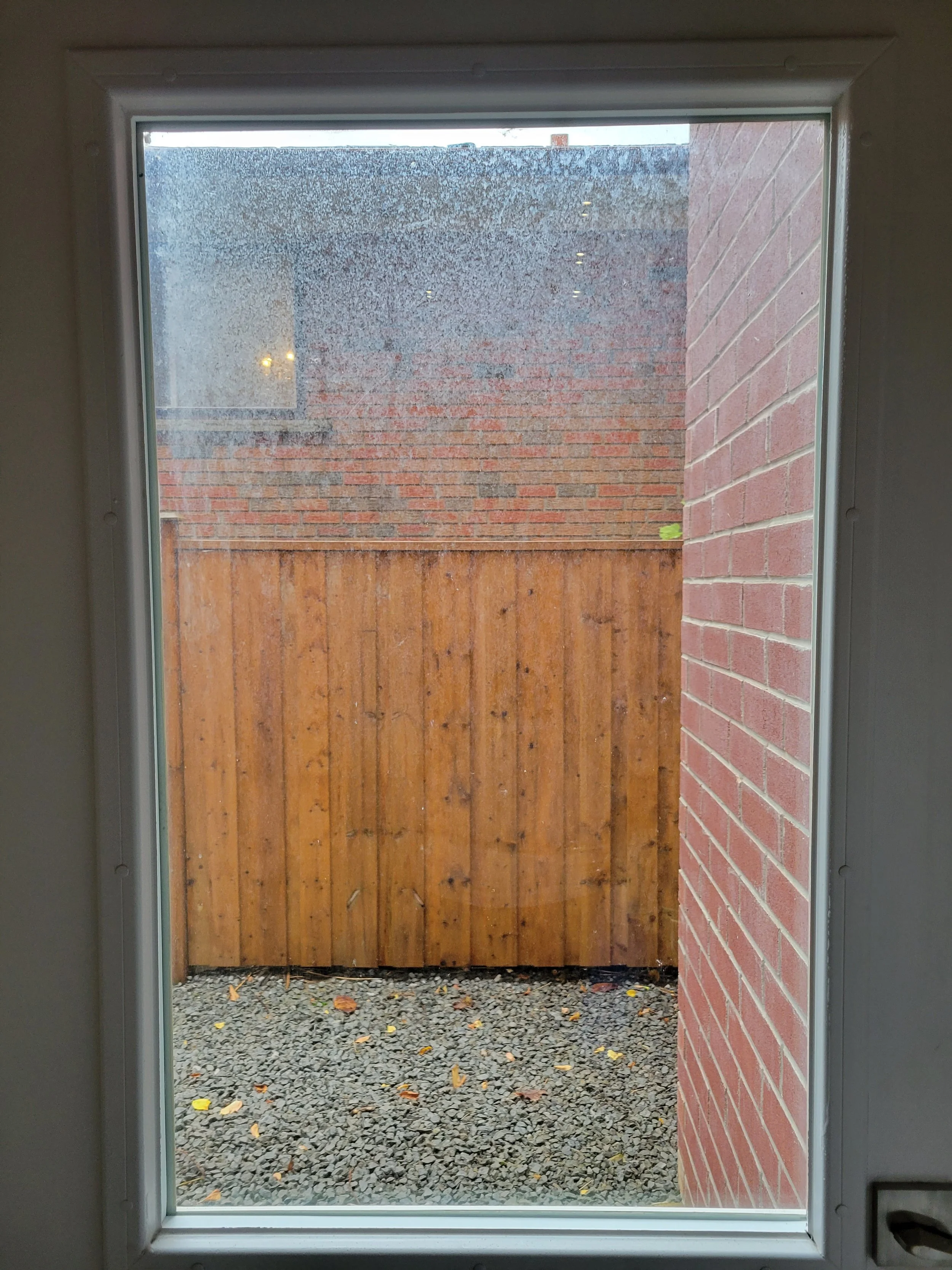 View through a window showing a small outdoor space with gravel ground, a wooden fence, and brick walls on each side, with rain visible on the window.