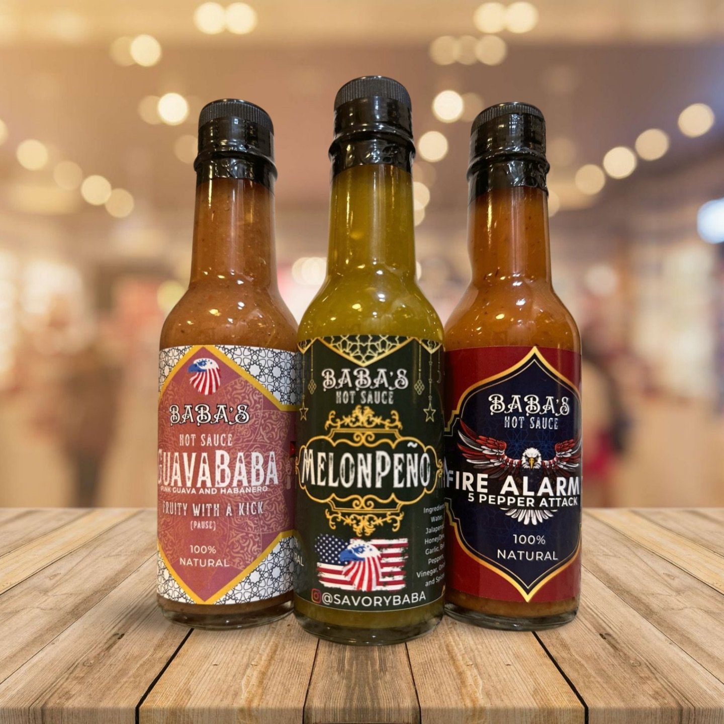 Three bottles of hot sauce on a wooden surface, with blurred background. The bottles are labeled "GuavaBaba," "MelonPeno," and "Fire Alarm."