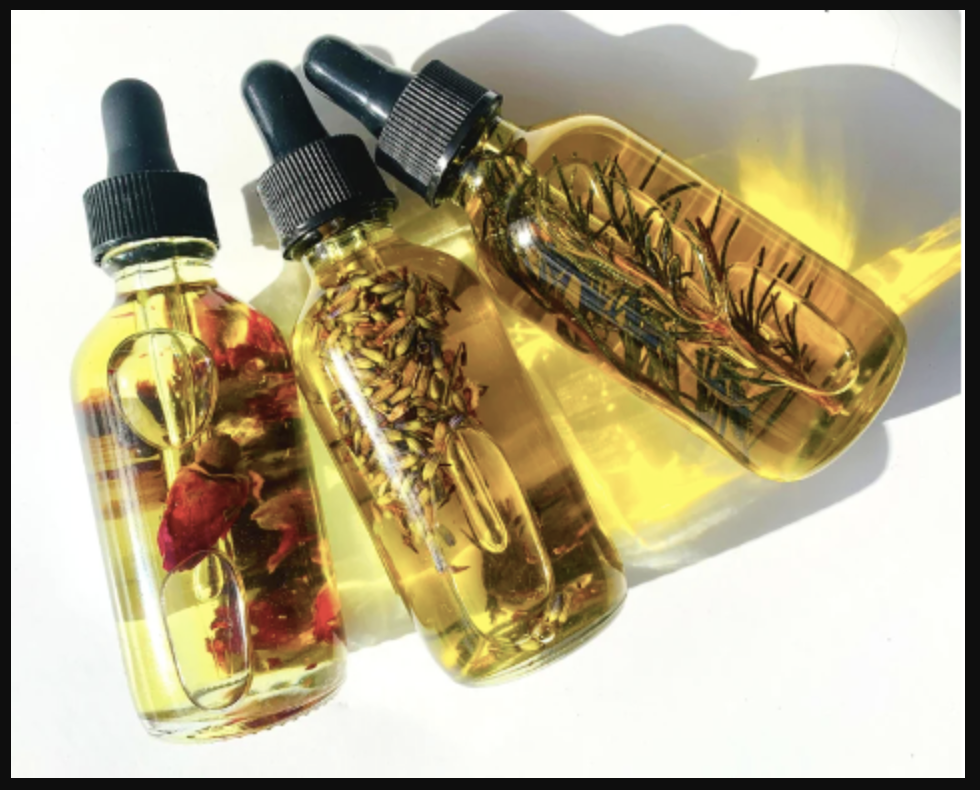 Three small glass bottles filled with herbal oil, each containing different dried herbs or flowers, with black dropper caps, grouped together on a white surface.