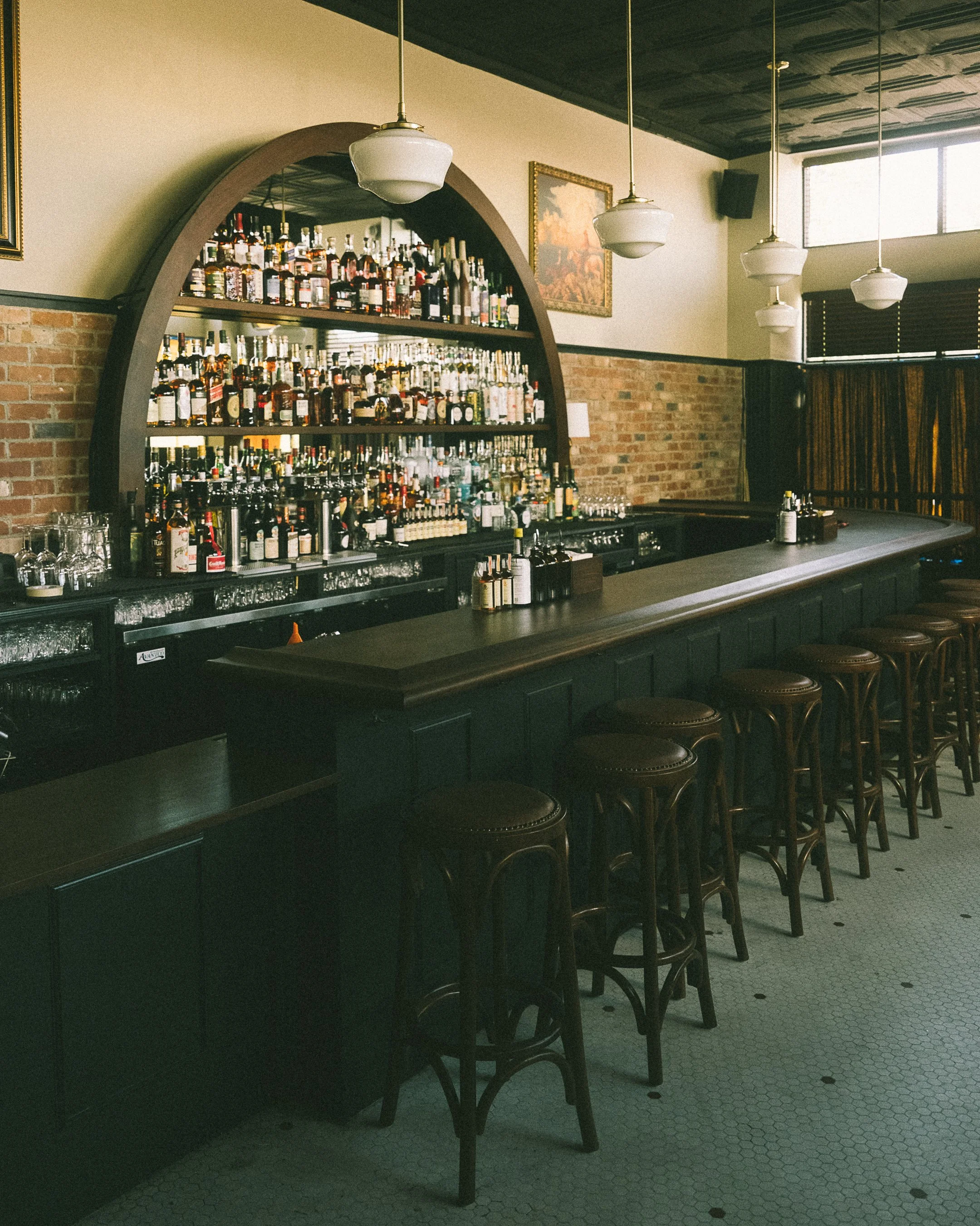 A long custom bar with liquor bottles on shelves behind the counter, bar stools along the counter, and a brick wall. Hanging white pendant lights, a framed picture, and large windows with blinds. Historic building.