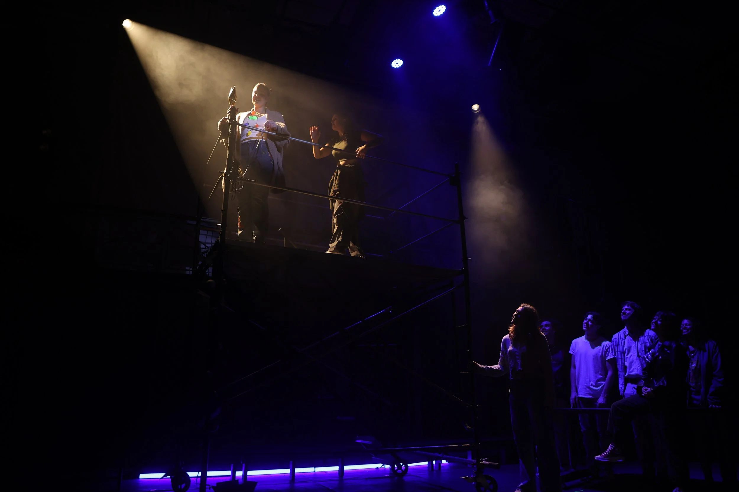 People sitting in a theater with a stage illuminated by spotlights, with two individuals performing or participating on the stage.