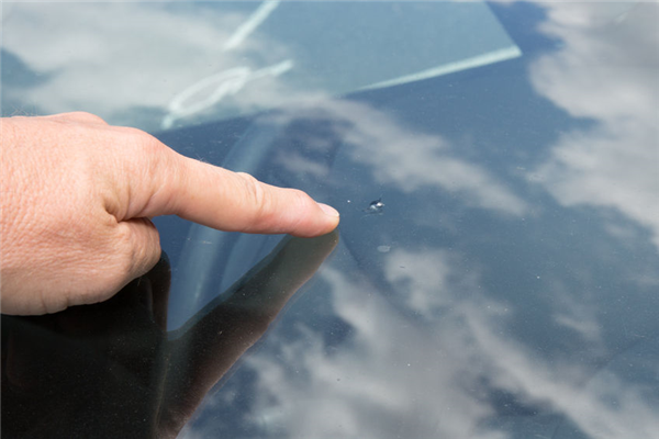 Close-up of a human finger pointing to a small chip on a car windshield with a cloudy sky reflection.