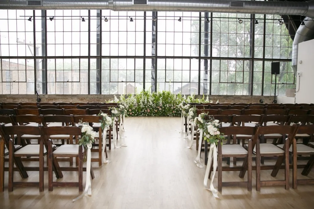 Outdoor wedding ceremony setup with rows of white chairs and floral arrangements on black stands facing an arch decorated with pink, white, and green flowers, against a red wooden barn wall.