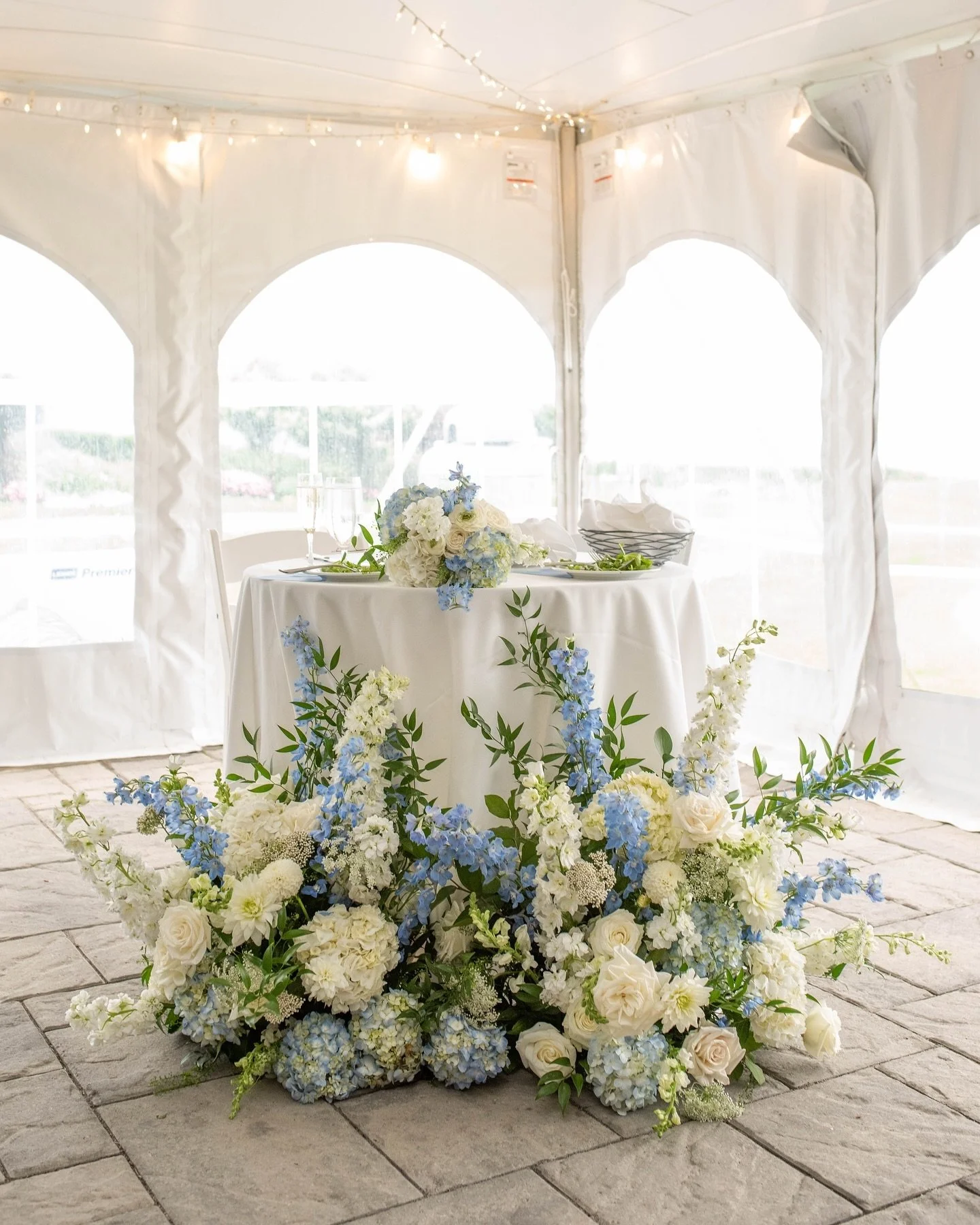 A woman in a white dress with black dots excitedly holding up a sheer veil, standing next to a man in a light gray suit holding a bouquet of baby's breath flowers covering his face, against a dark wooden wall background.