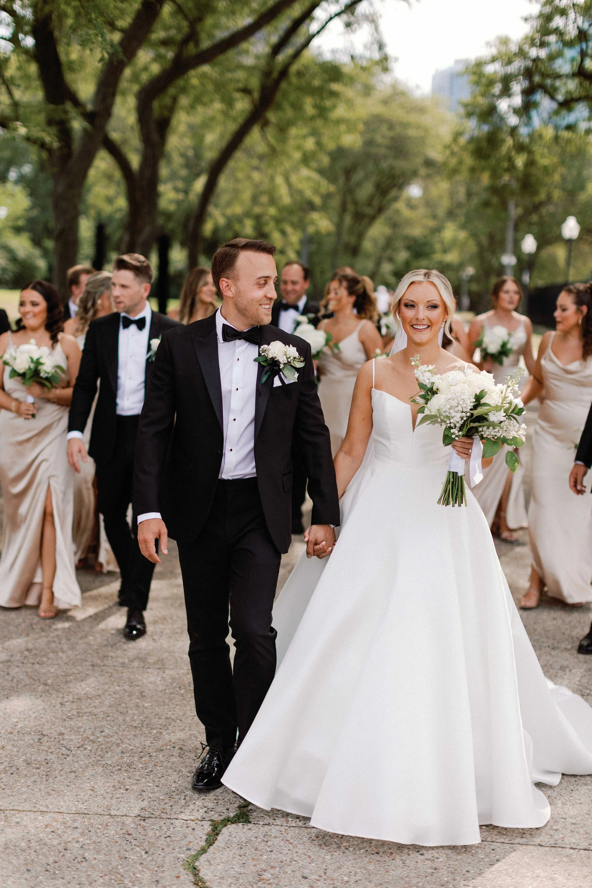 A couple dressed in wedding attire smiling and embracing outdoors in a natural setting with greenery.