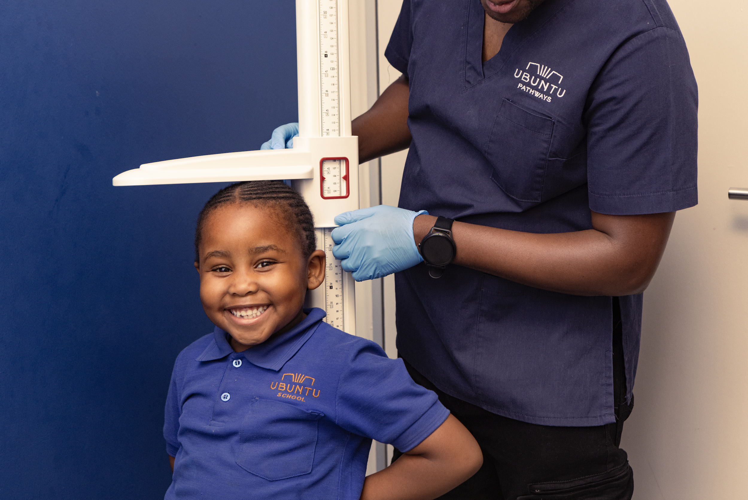 A smiling young boy in a blue polo shirt standing in front of a blue wall and measuring himself against a height scale with a medical professional adjusting the scale. The professional is wearing a navy blue shirt with the logo 'Ubuntu Pathways' and 