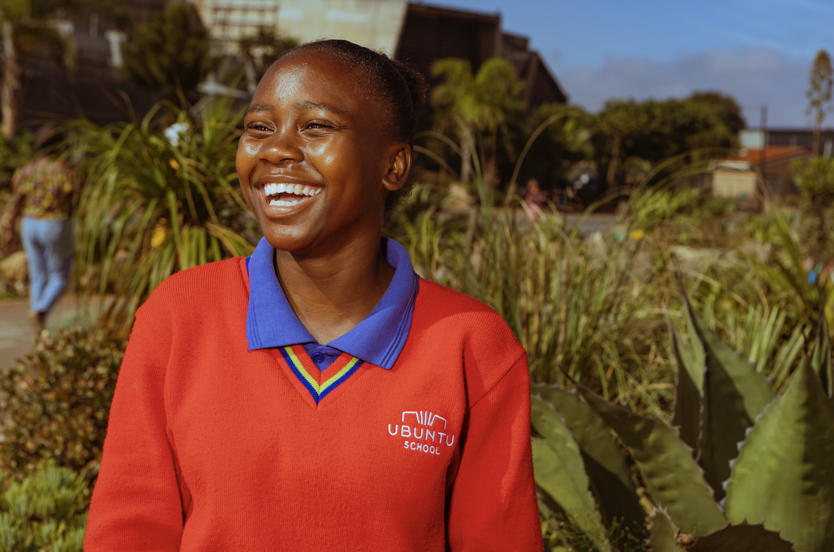 A young girl with a bright smile, wearing a red school uniform with a blue collar, standing outdoors with lush green plants and buildings in the background.