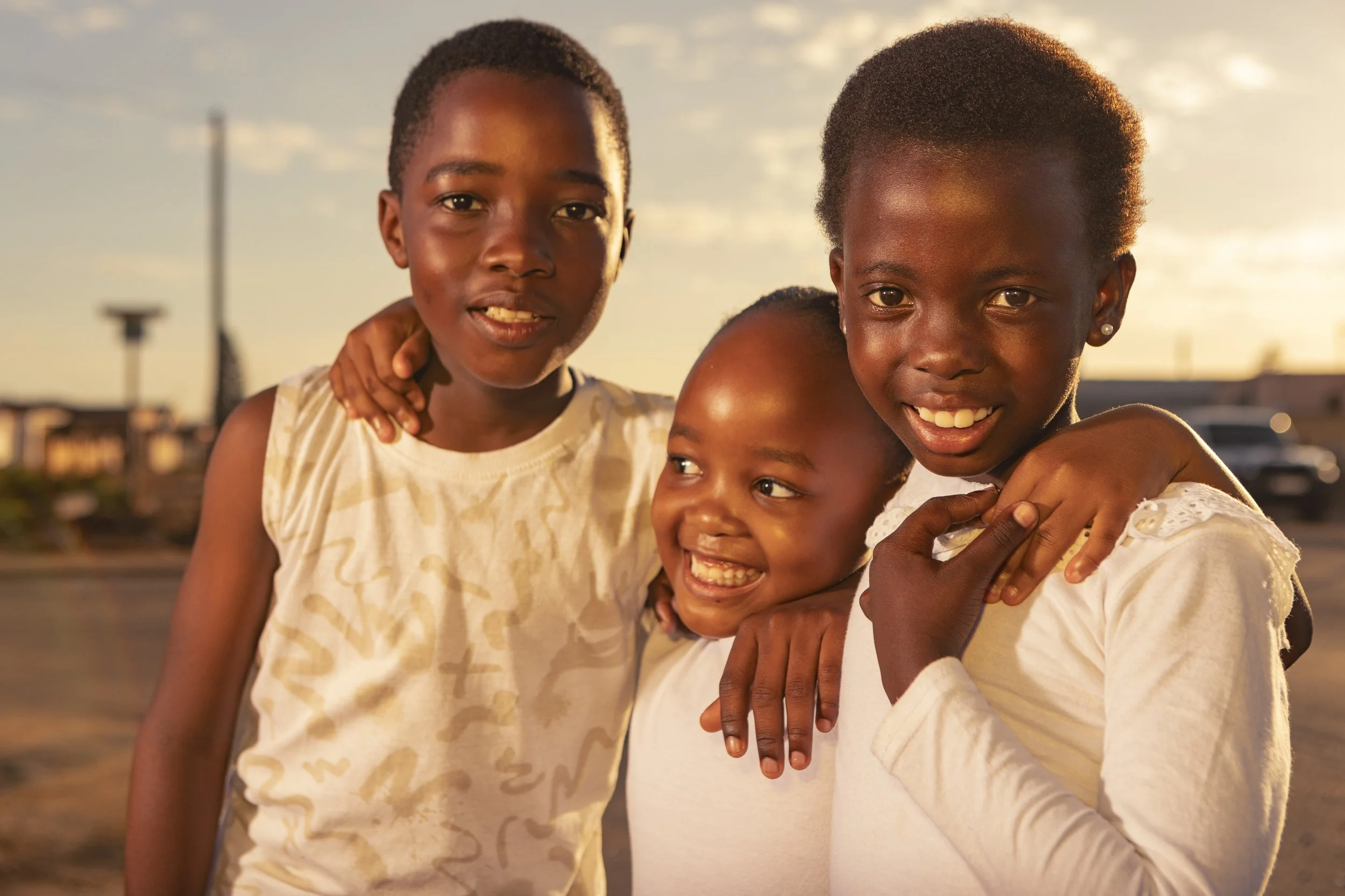 Three smiling children, two girls and one boy, embrace each other outside during sunset.