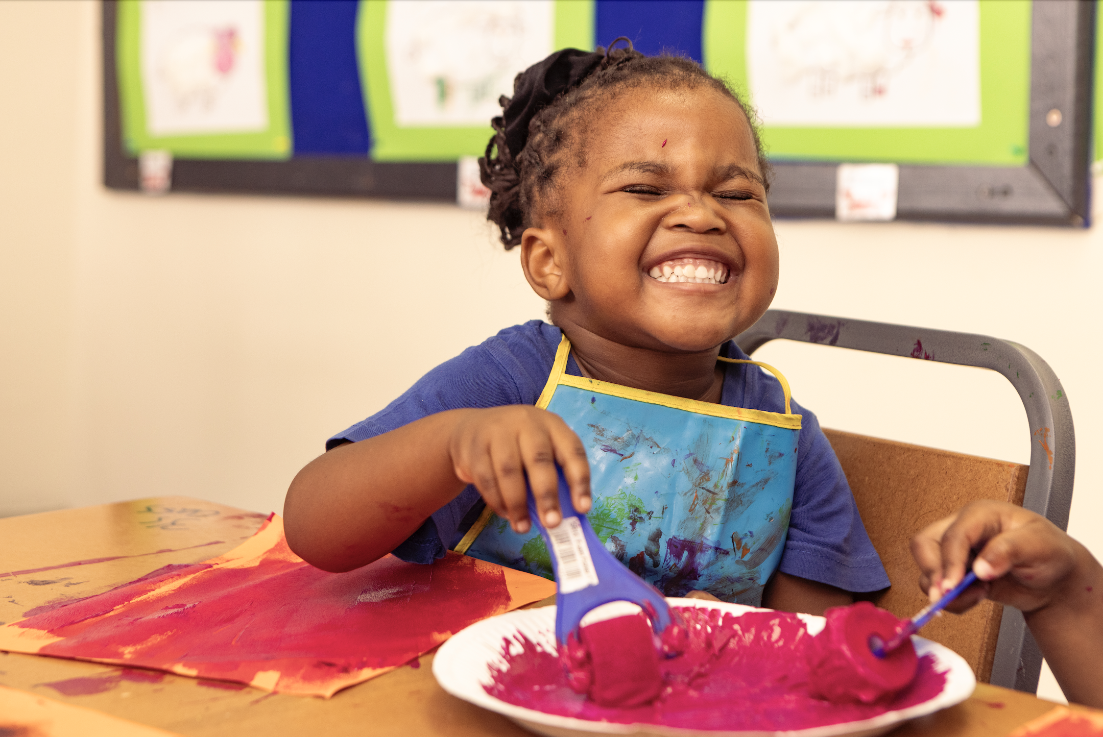 A young girl with dreadlocks and a big smile sits at a table covered with colorful art supplies, including pink paint, scissors, and paper with painted patterns. She wears a blue shirt, a paint-spattered apron, and is engaging in a painting activity 