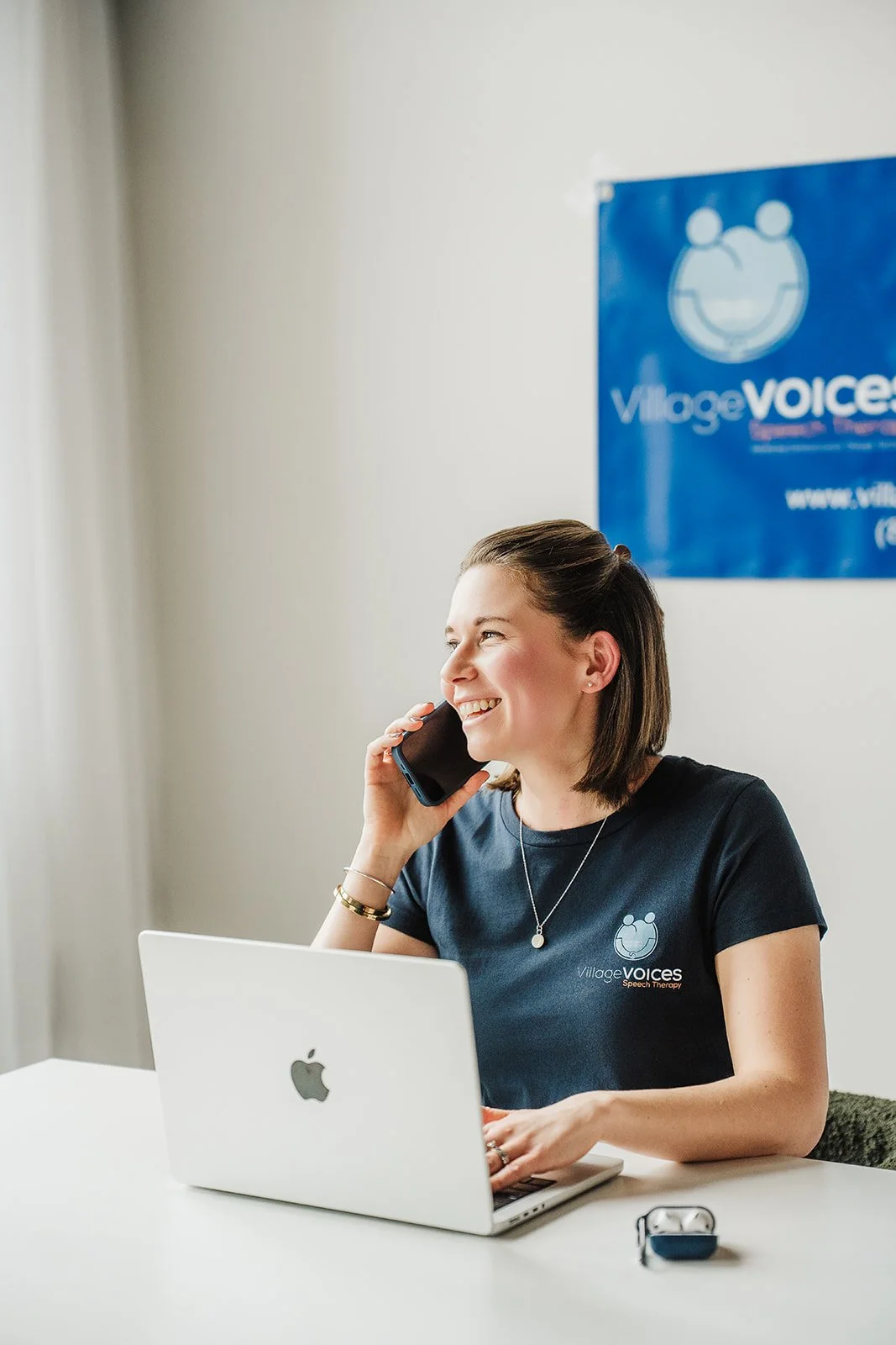 A woman smiling while sitting in front of a laptop and holding a phone up to her right ear.